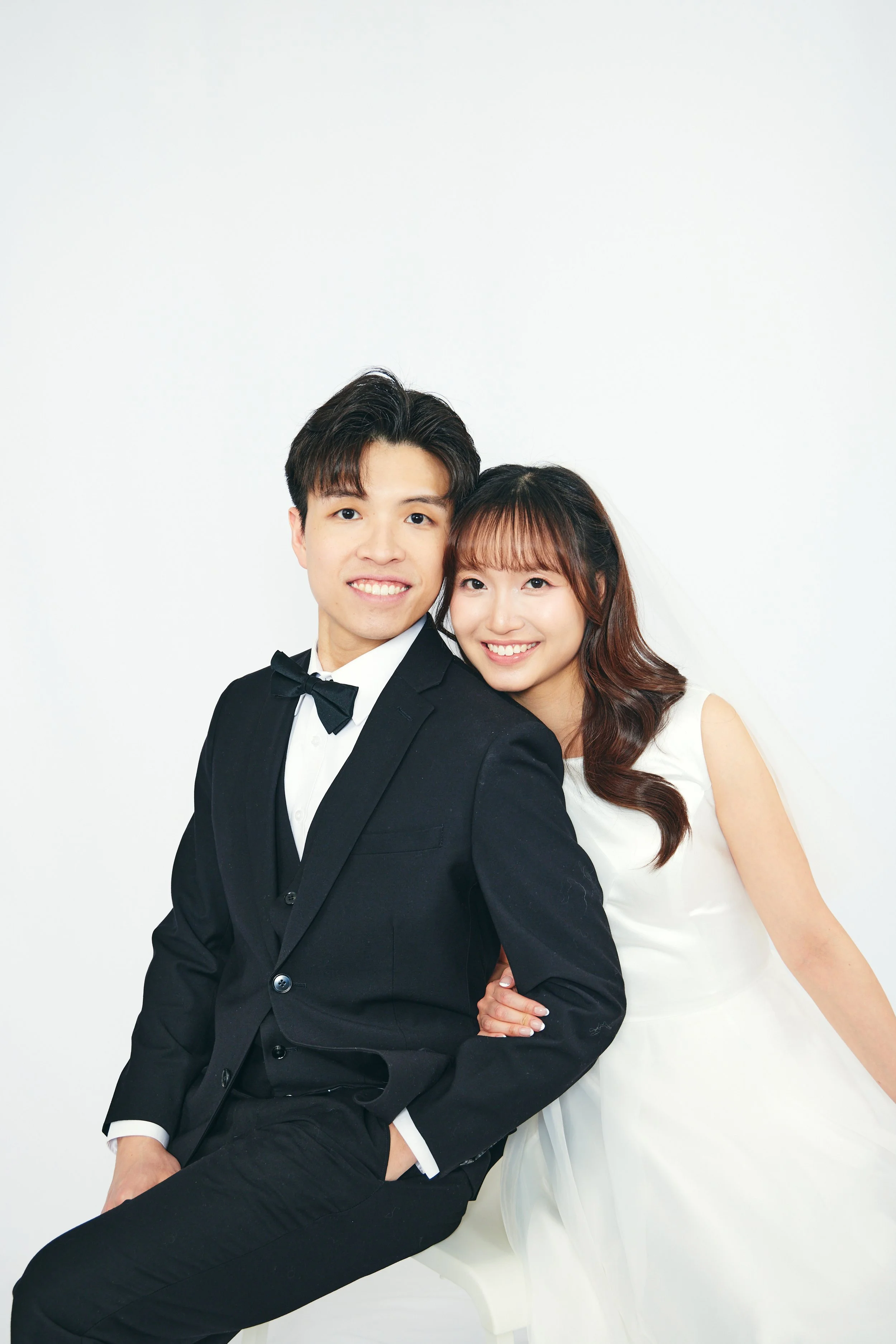 Young couple dressed in formal wedding attire, smiling and posing against a plain white background. The man is wearing a black tuxedo with a bow tie, and the woman is wearing a white wedding dress.
