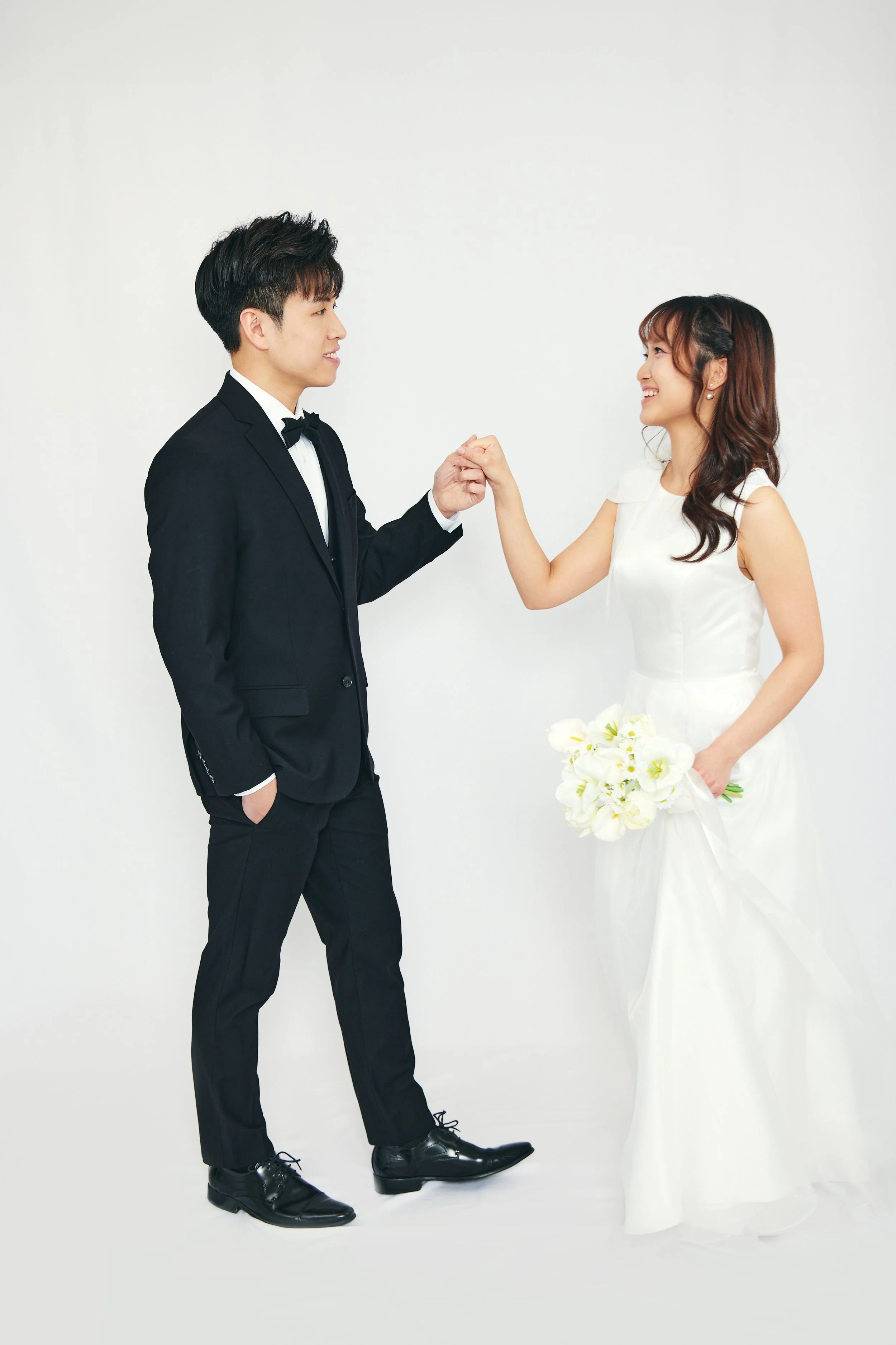 A bride and groom in wedding attire holding hands and smiling at each other against a plain white background.