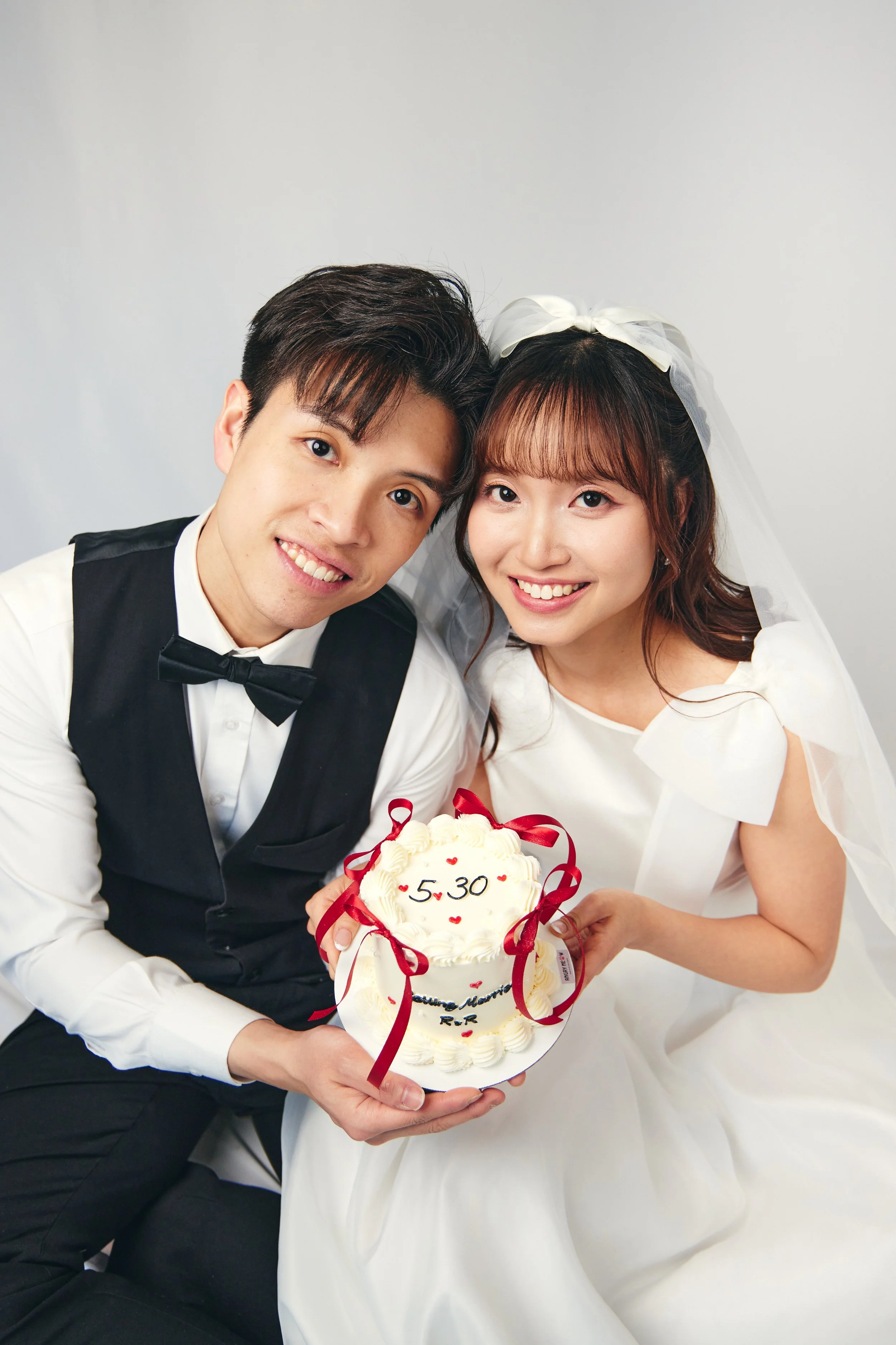 A happy couple dressed in wedding attire holding a wedding cake with the date 5.30 written on it, smiling at the camera against a plain background.