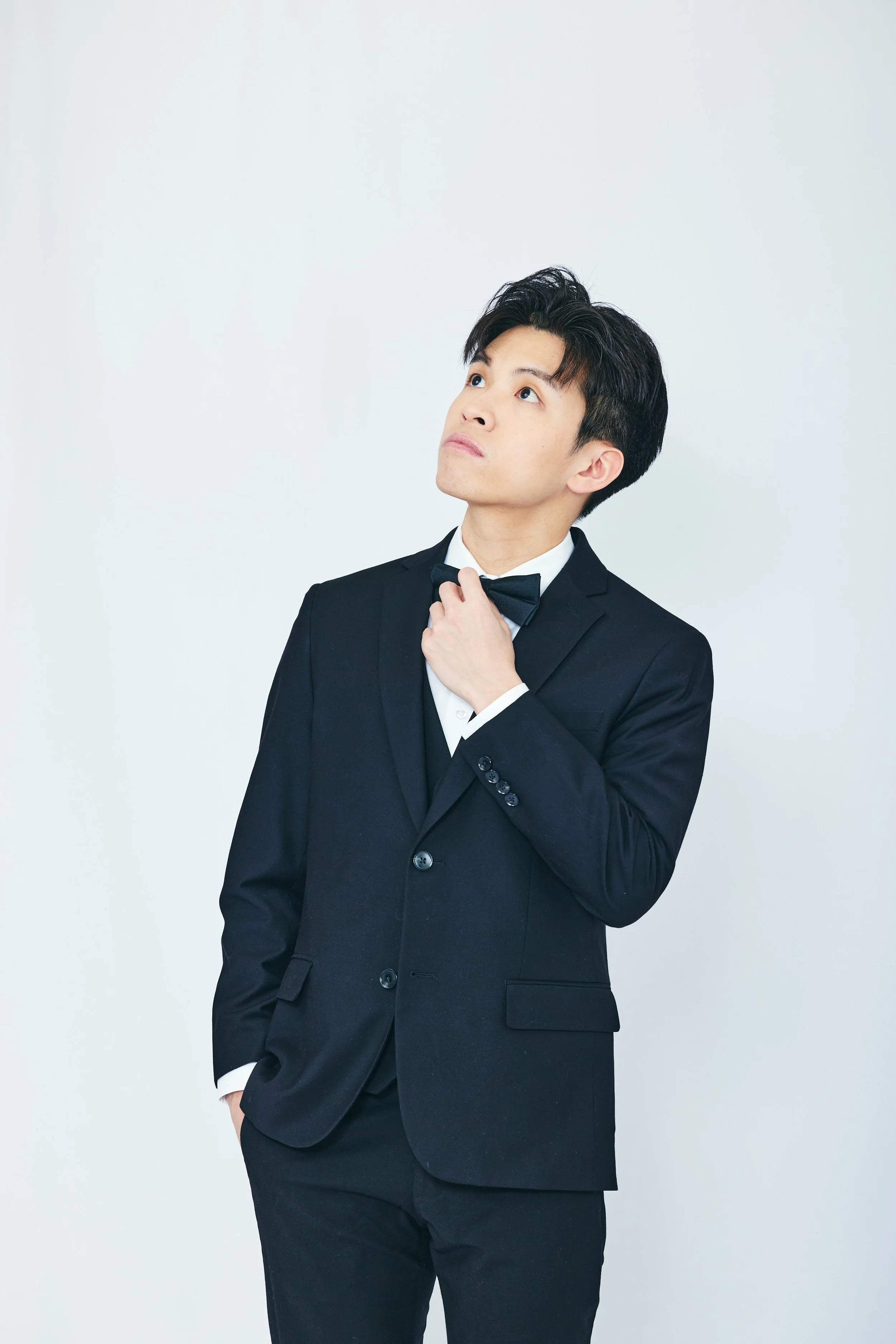 A young man in a black tuxedo adjusting his bow tie, standing against a plain white background.