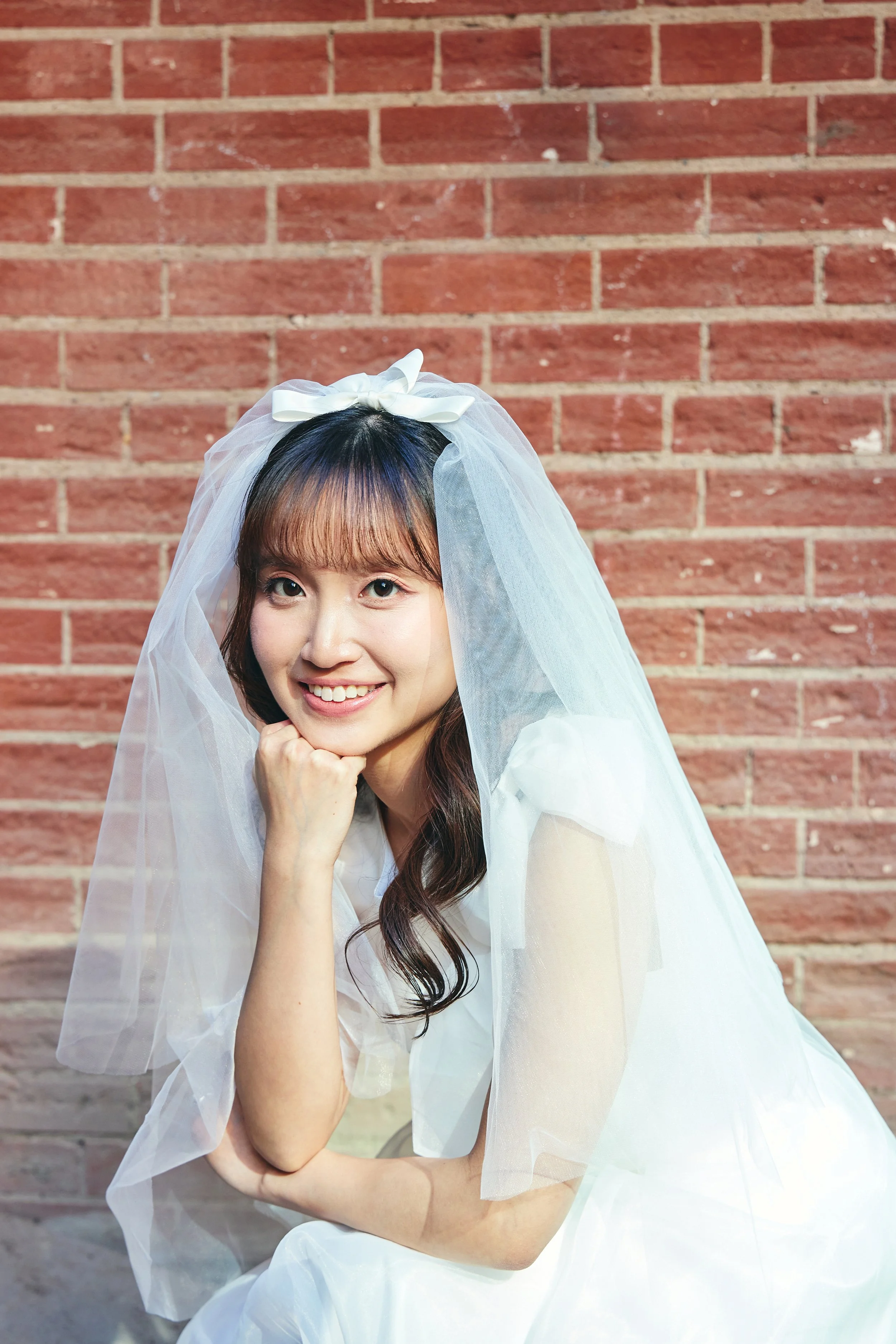 A young woman with long brown hair and bangs, dressed in a white dress and veil with a bow, smiling and resting her chin on her hand in front of a red brick wall.