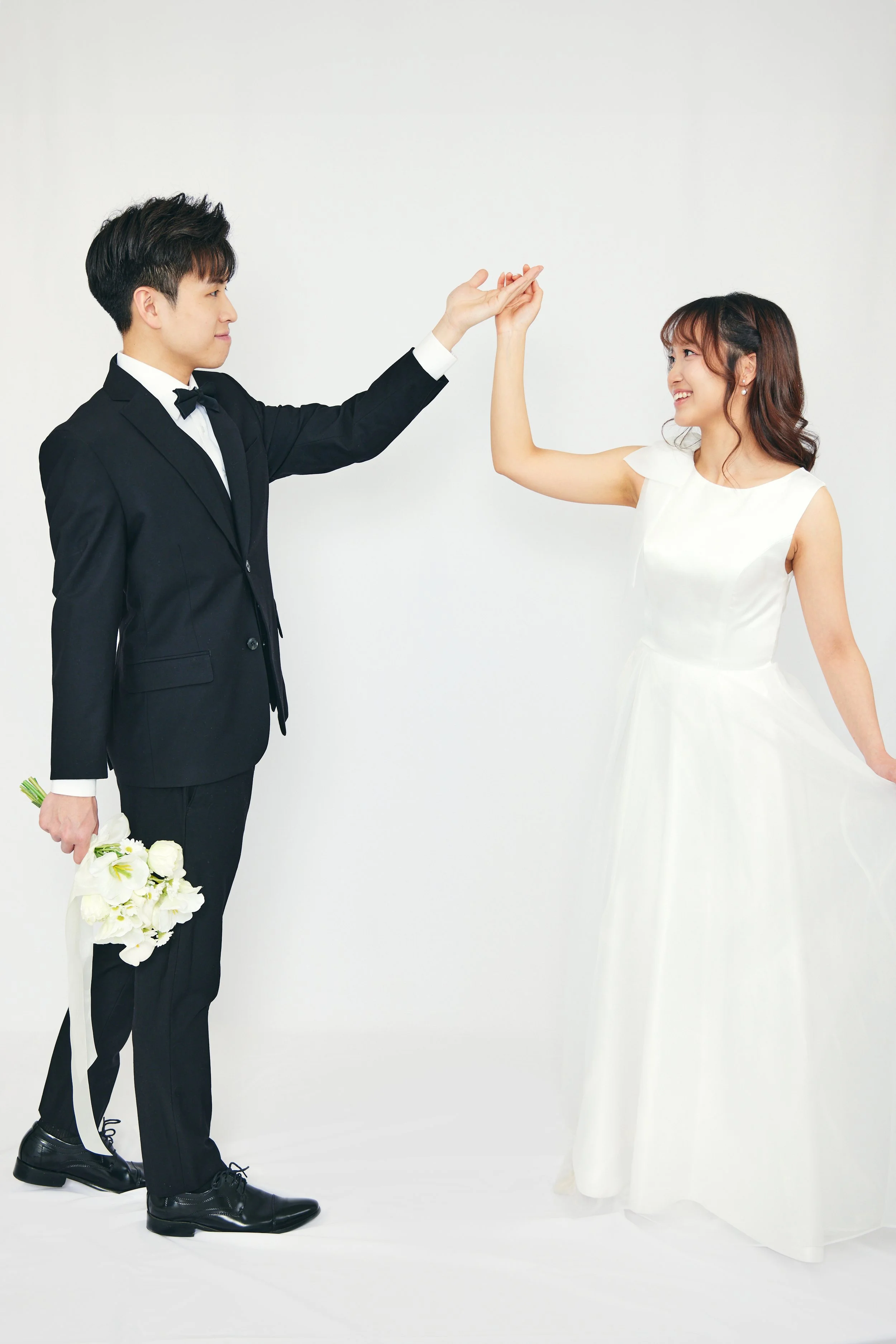 A man in a black tuxedo holding a bouquet of white flowers with a white ribbon, and a woman in a white wedding dress, are standing against a plain white background. The woman is smiling and raising her hand toward the man's hand as they prepare for a