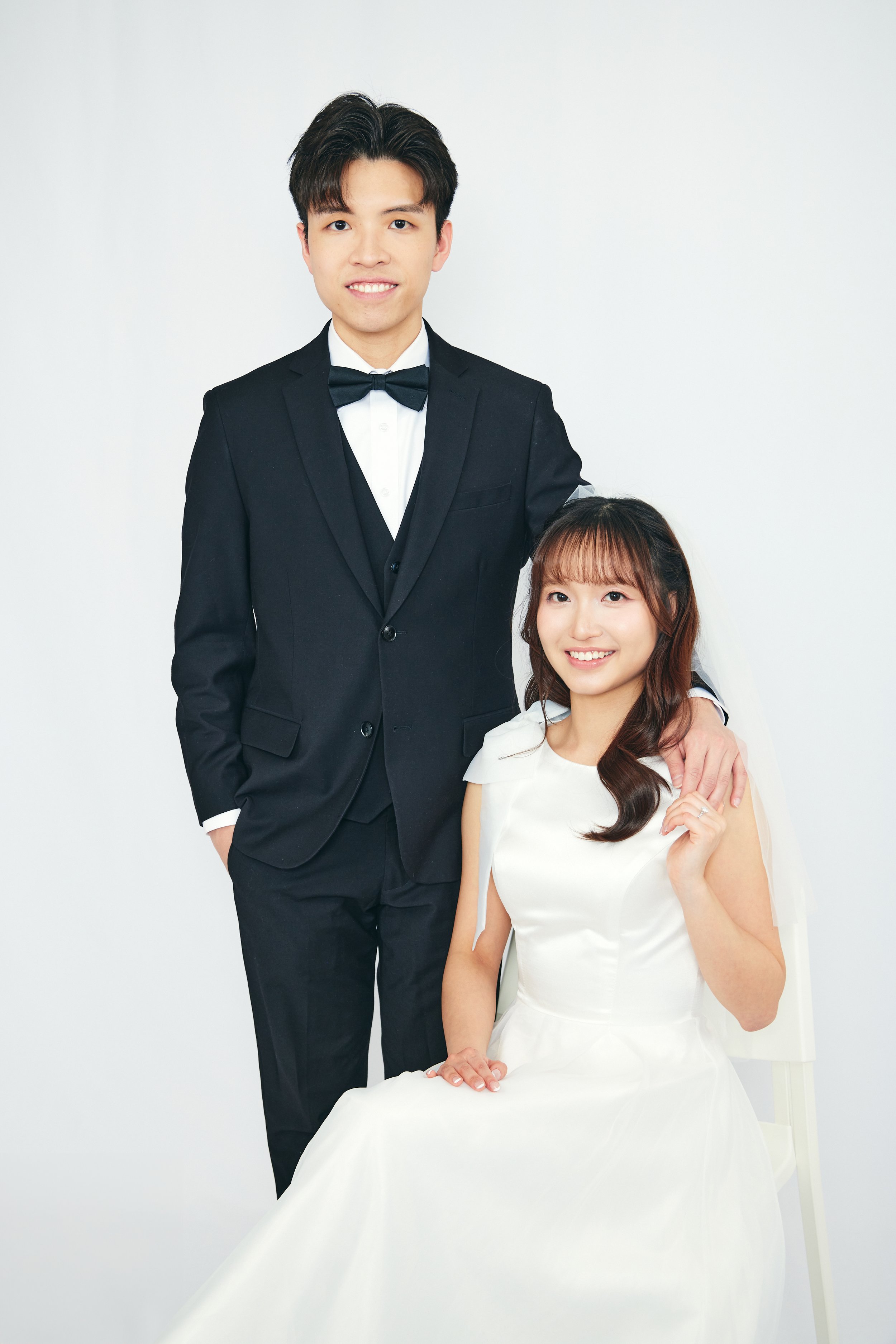 A young man in a black tuxedo standing with his arm on the shoulder of a young woman in a white wedding dress who is sitting and smiling.