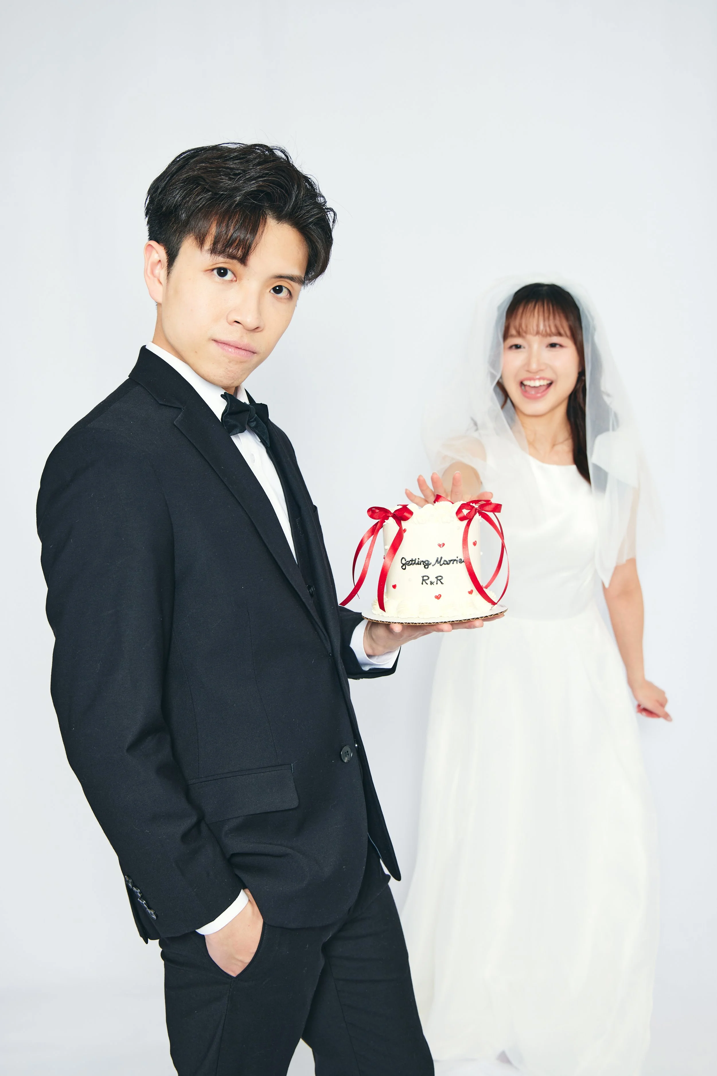 A groom in a black tuxedo holding a wedding cake with a bride in a white wedding dress and veil smiling in the background.