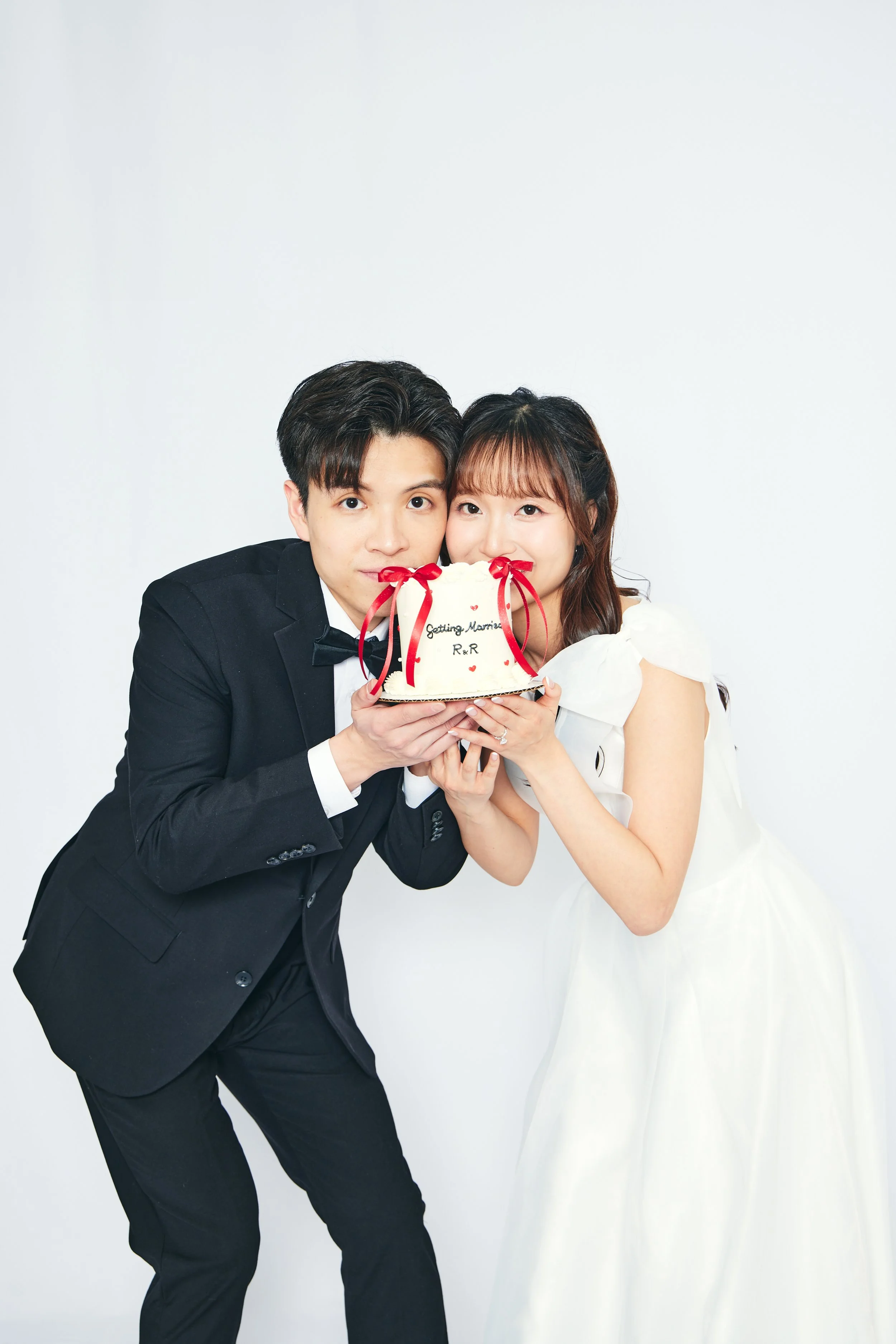 A couple dressed in wedding attire holding a small white wedding cake with red ribbons, celebrating their wedding with a plain white background.