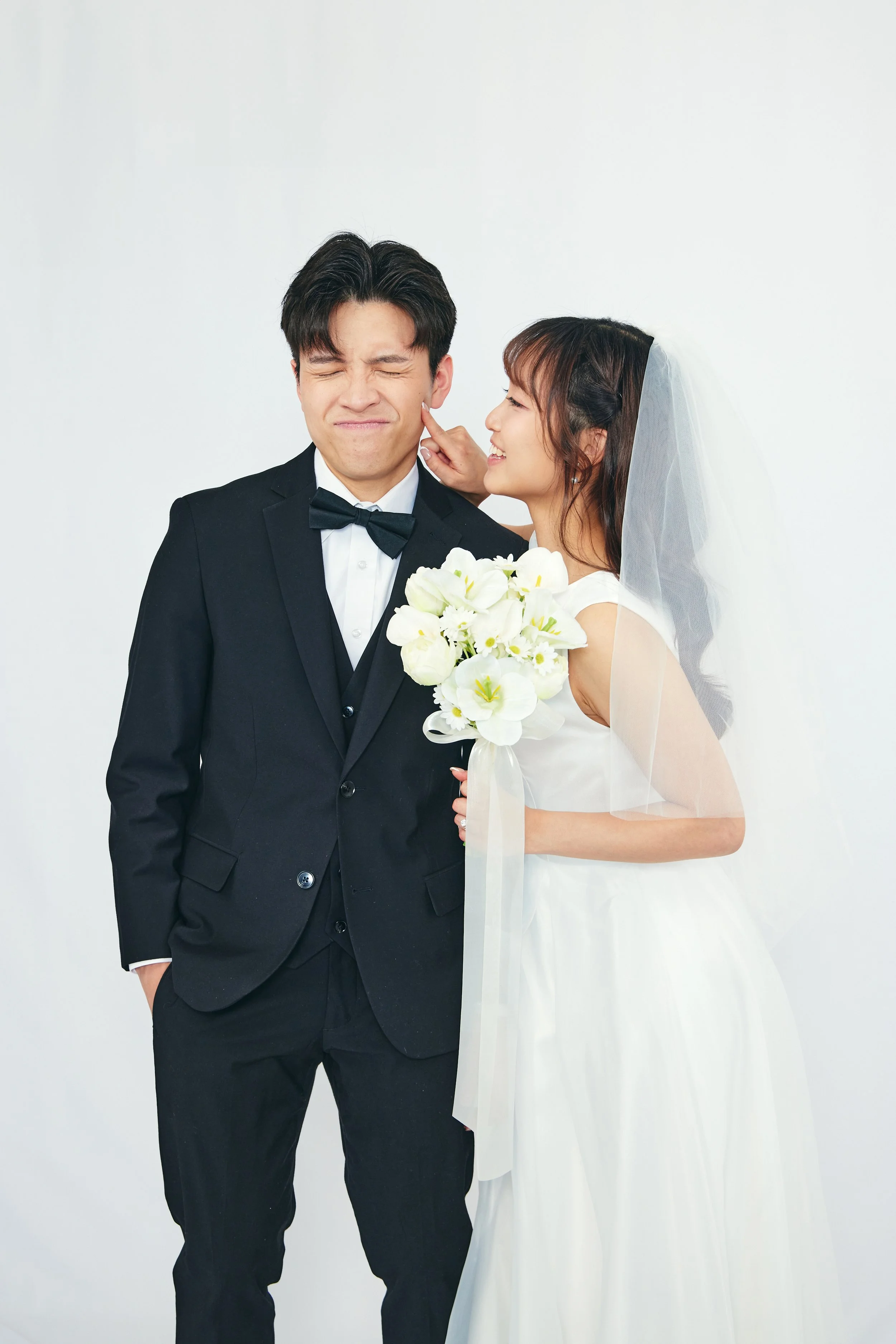 A bride and groom are in a photo. The groom is in a black tuxedo with a bow tie, and the bride is in a white wedding dress with a veil and holding a bouquet of white flowers. The bride is touching the groom's cheek, and he looks happy and laughing.