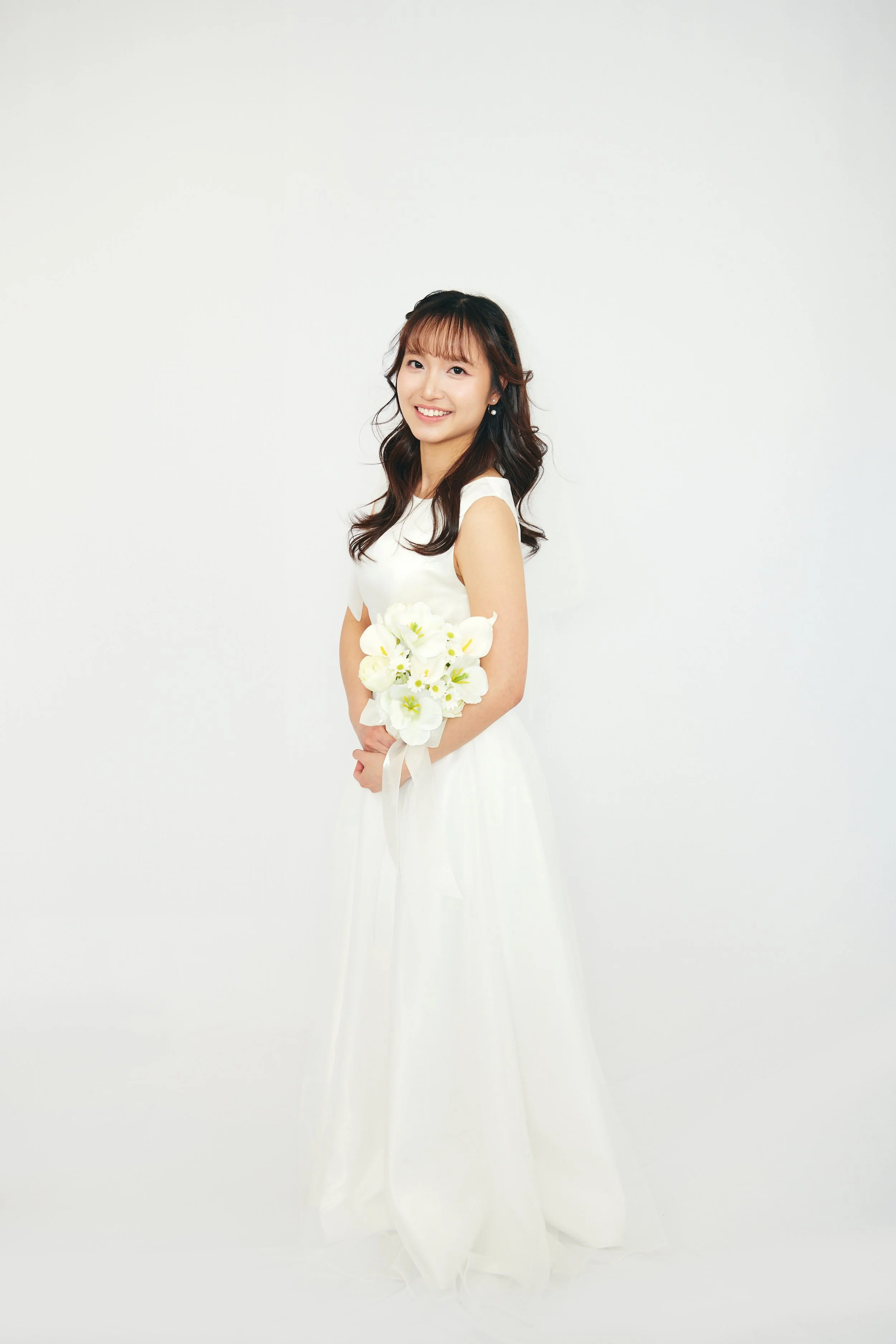 Young woman in white wedding dress holding a bouquet of white flowers, smiling against a plain white background.