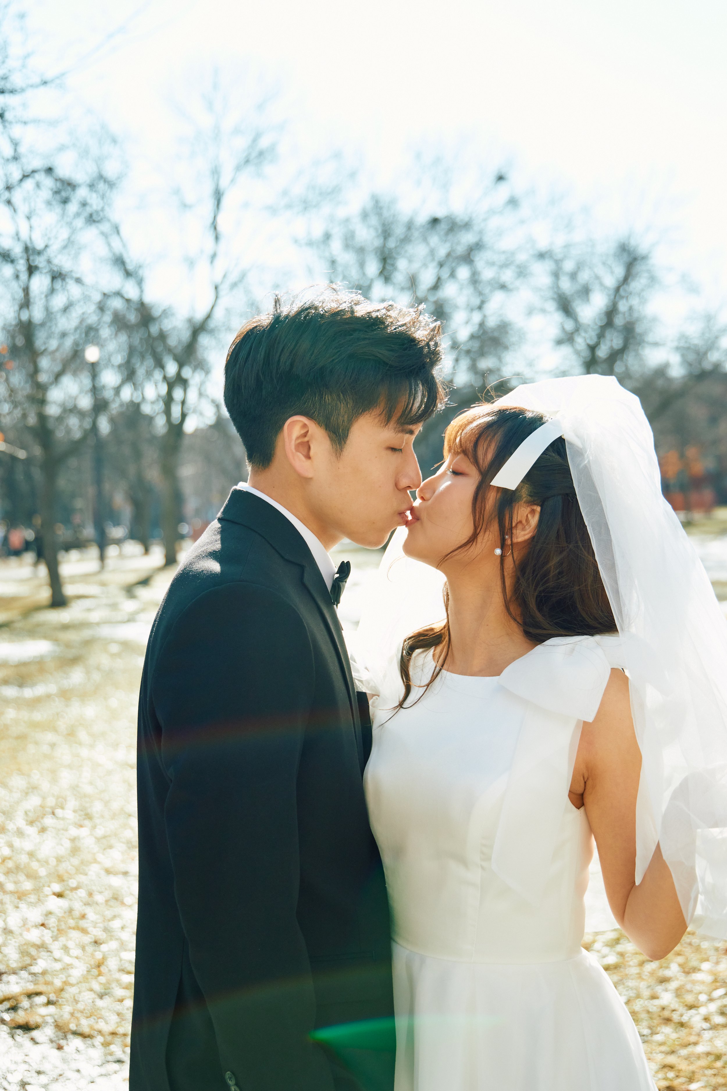 A couple about to kiss outdoors on a sunny day, with trees in the background. The man is in a black tuxedo, and the woman is in a white wedding dress with a veil.
