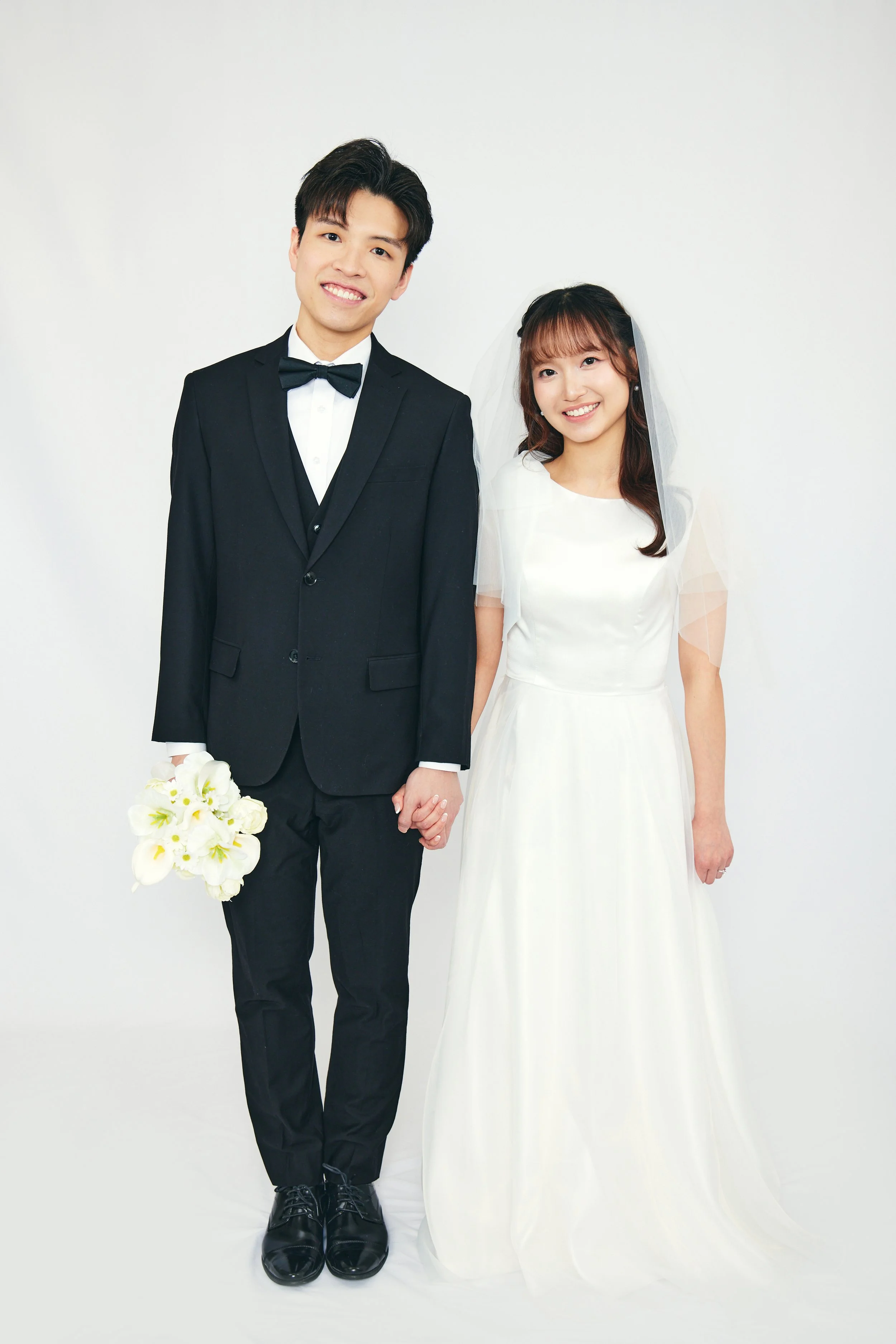 A bride and groom standing together, holding hands, smiling for a wedding photo. The groom is wearing a black tuxedo with a bow tie, and the bride is wearing a white wedding dress with a veil, holding a bouquet of white flowers.