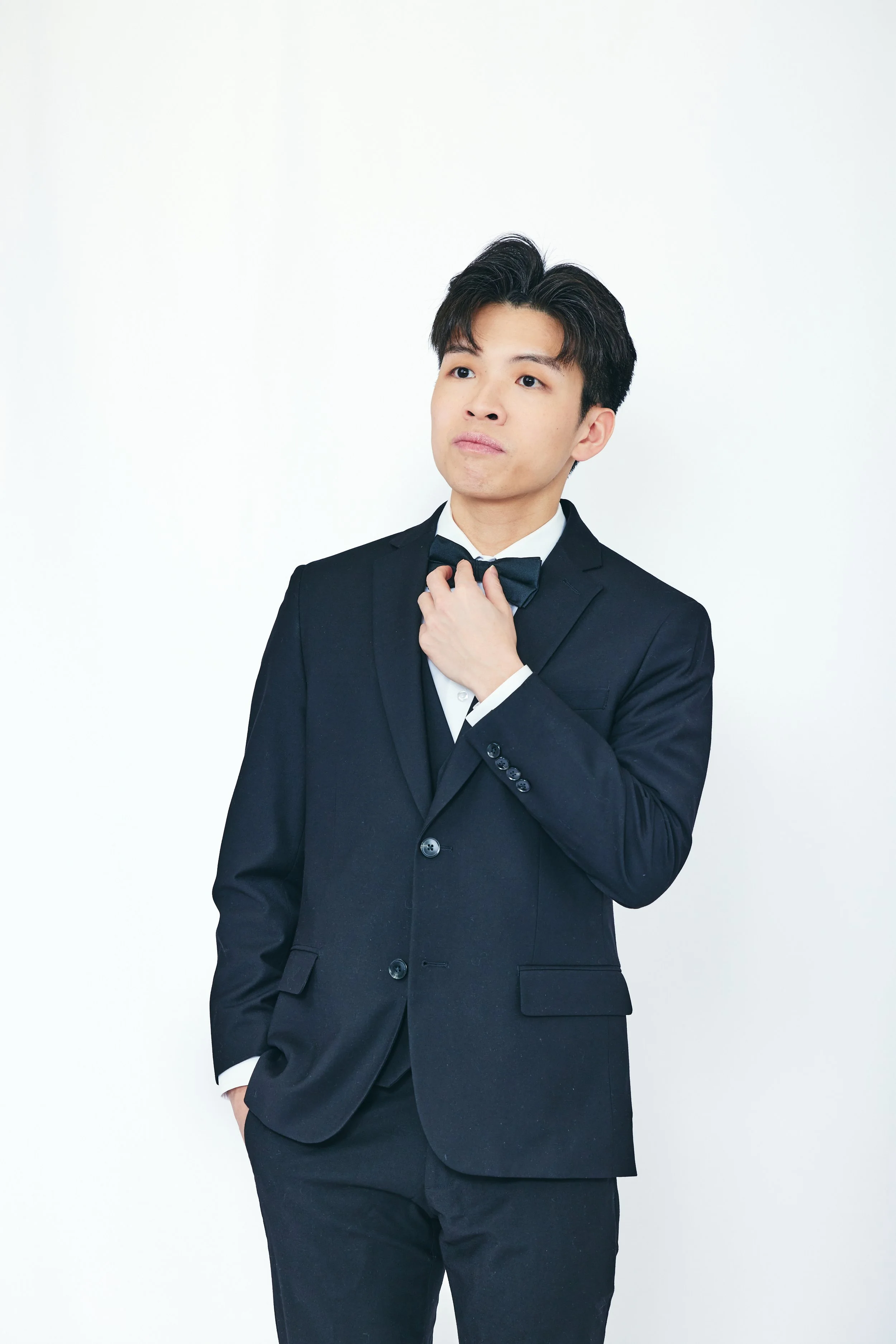 Young man in black tuxedo adjusting bow tie against plain white background.