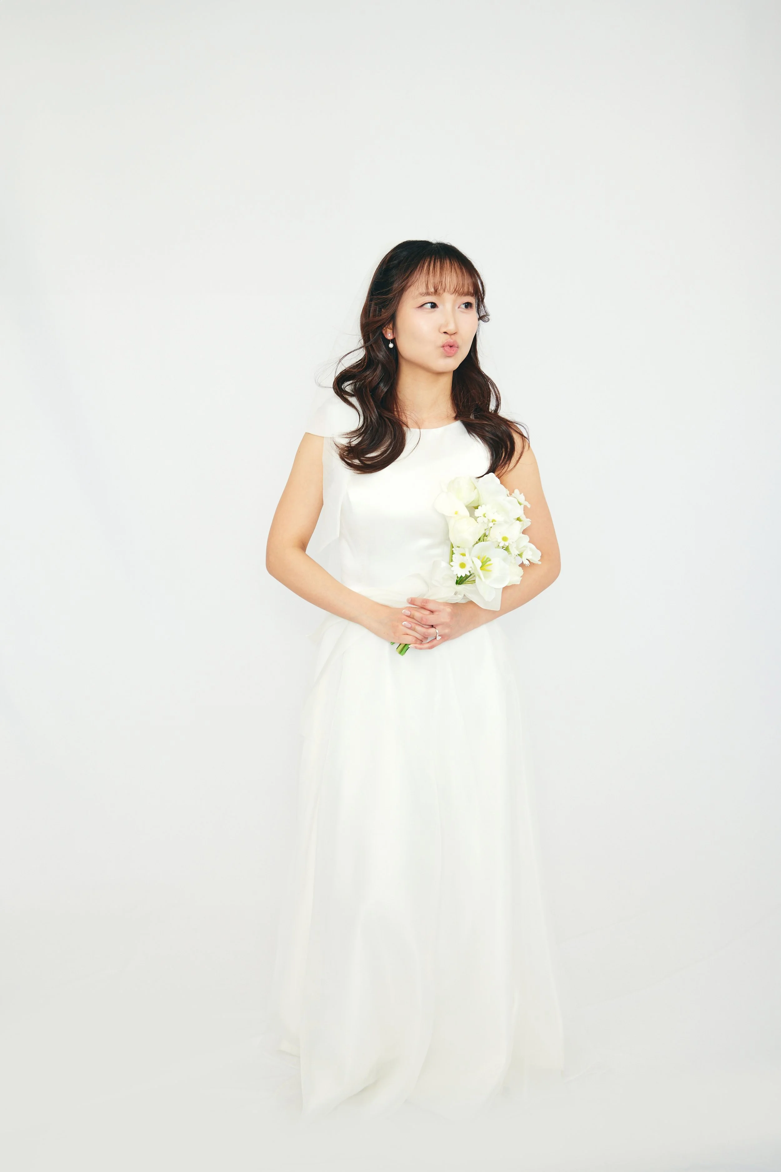 Young woman in a white dress holding a bouquet of white flowers, making a playful expression against a plain white background.
