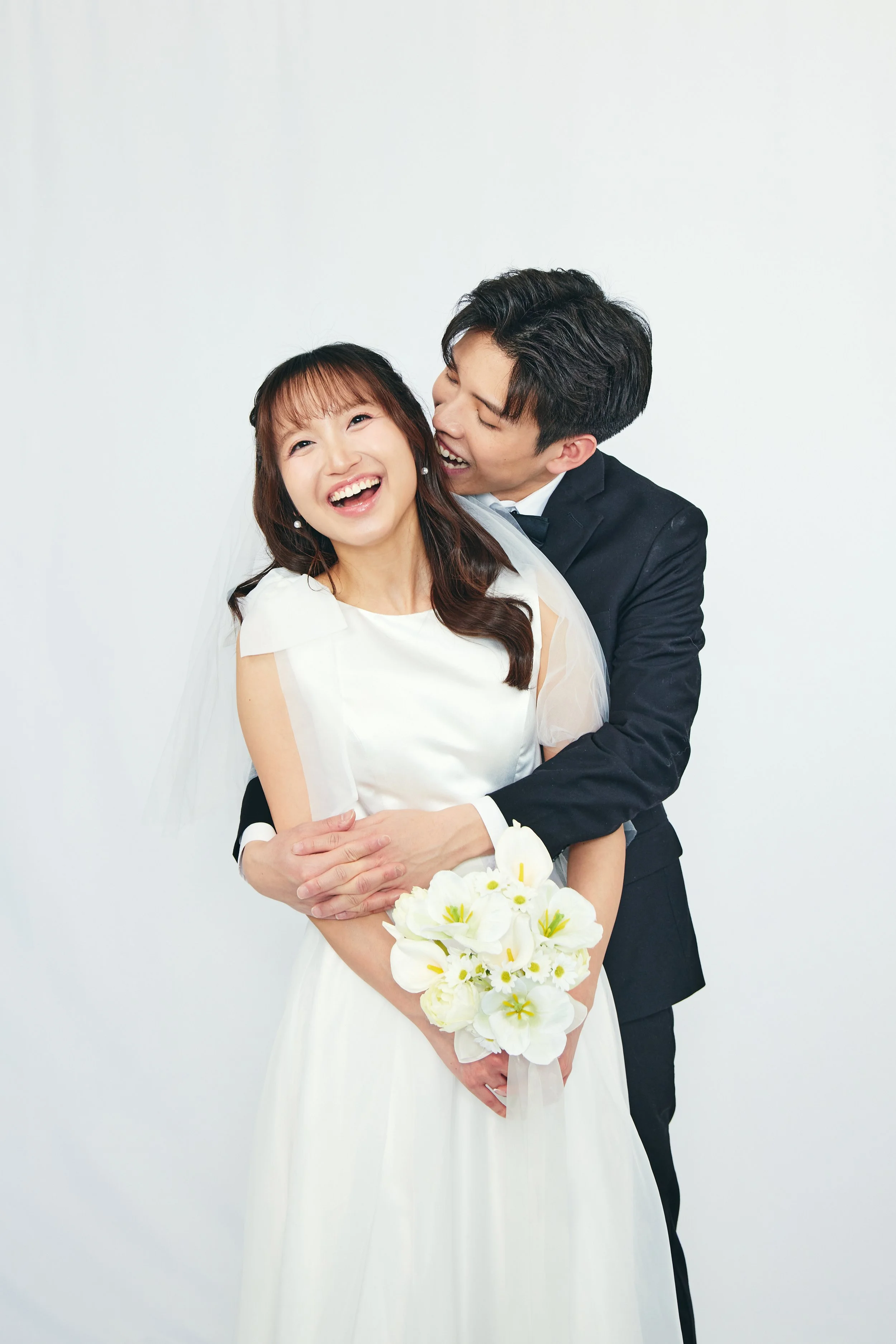 A joyful bride and groom are smiling and embracing, with the bride holding a bouquet of white flowers against a plain white background.