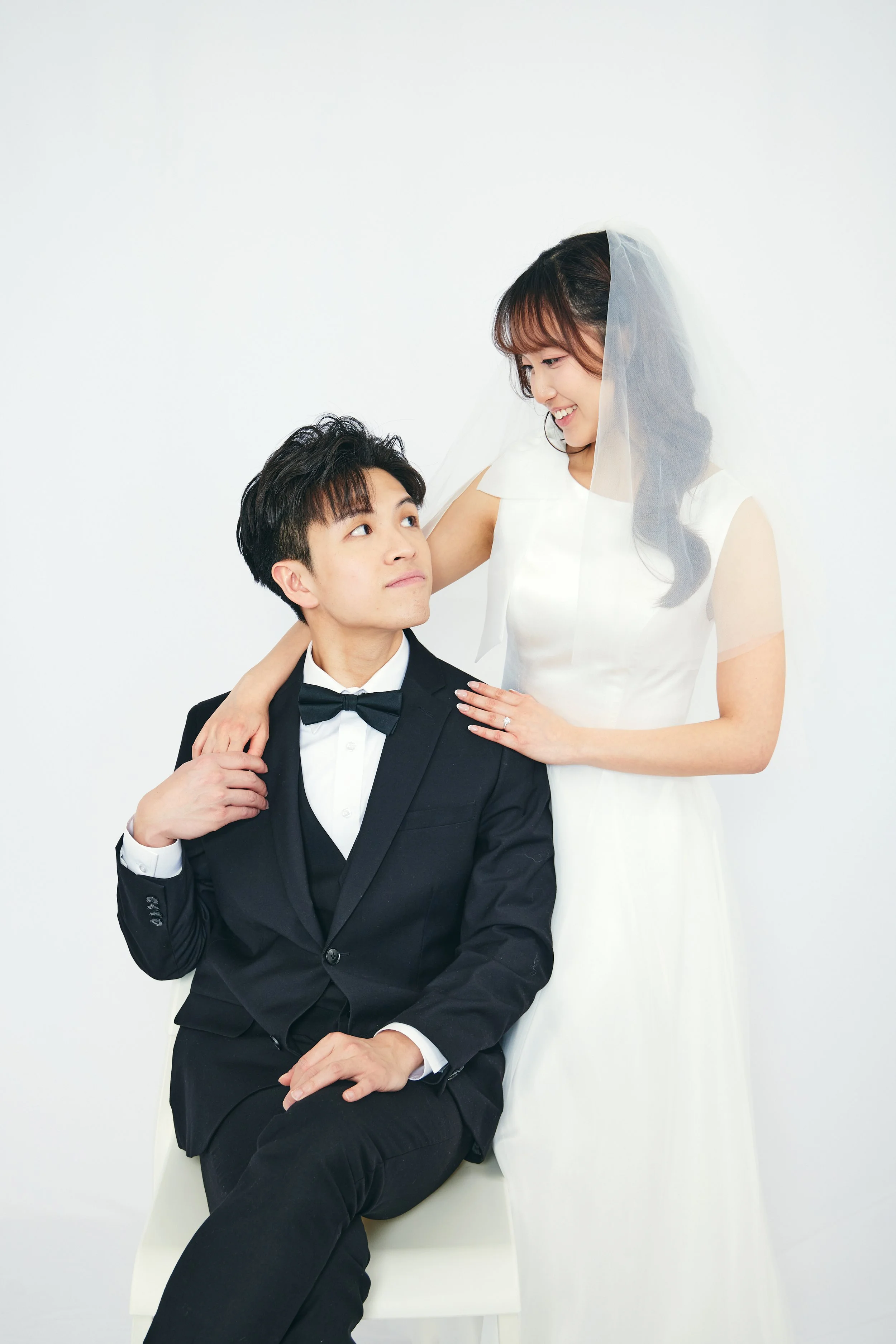 A bride in a white wedding dress and veil looks at a groom in a black tuxedo and bow tie, who is seated on a chair, against a plain white background.