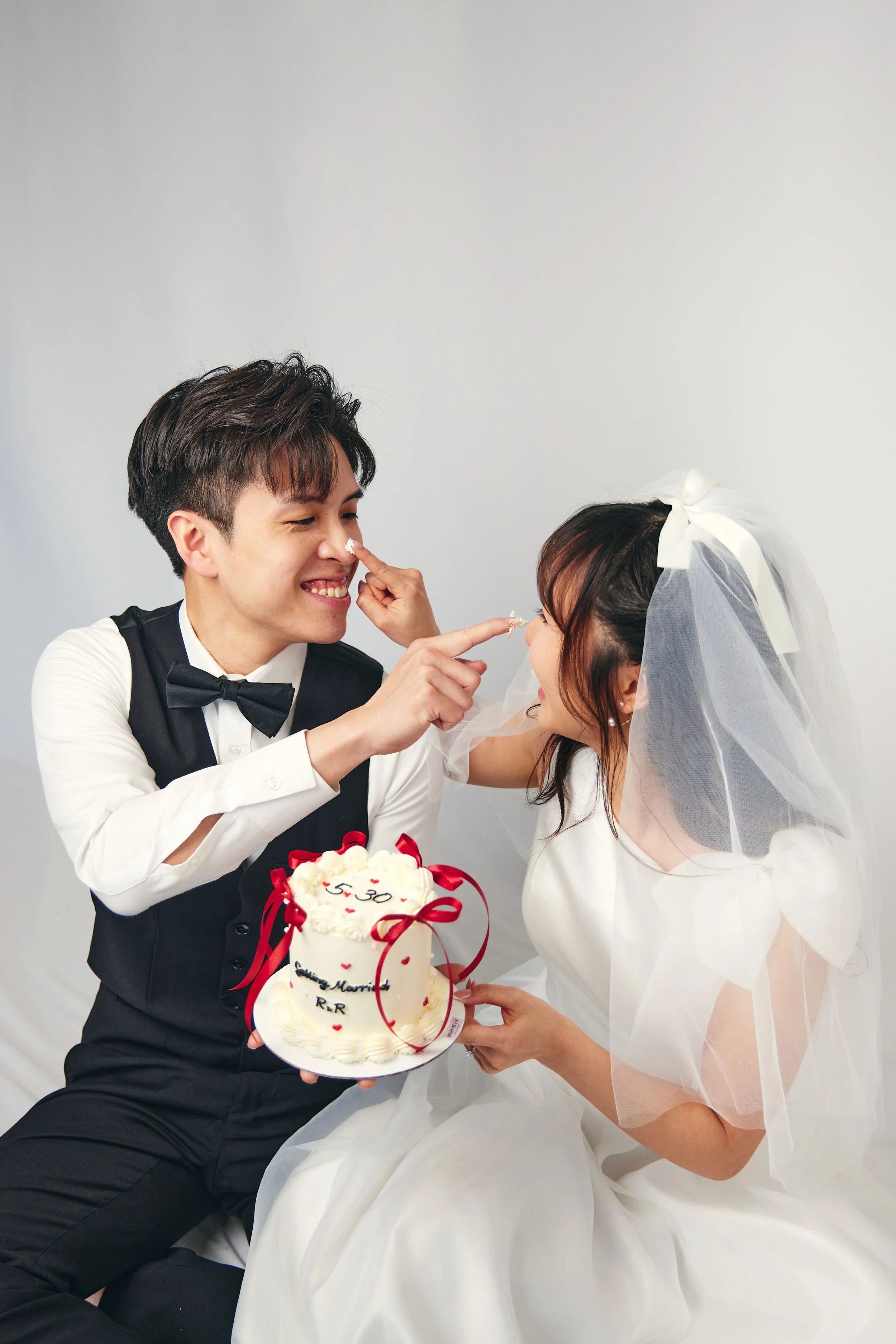 A newlywed couple smiling and playfully touching noses during their wedding celebration. The bride, in a white wedding dress with a veil, holds a wedding cake decorated with red ribbons and hearts. The groom, in a black tuxedo with a bow tie, smiles 