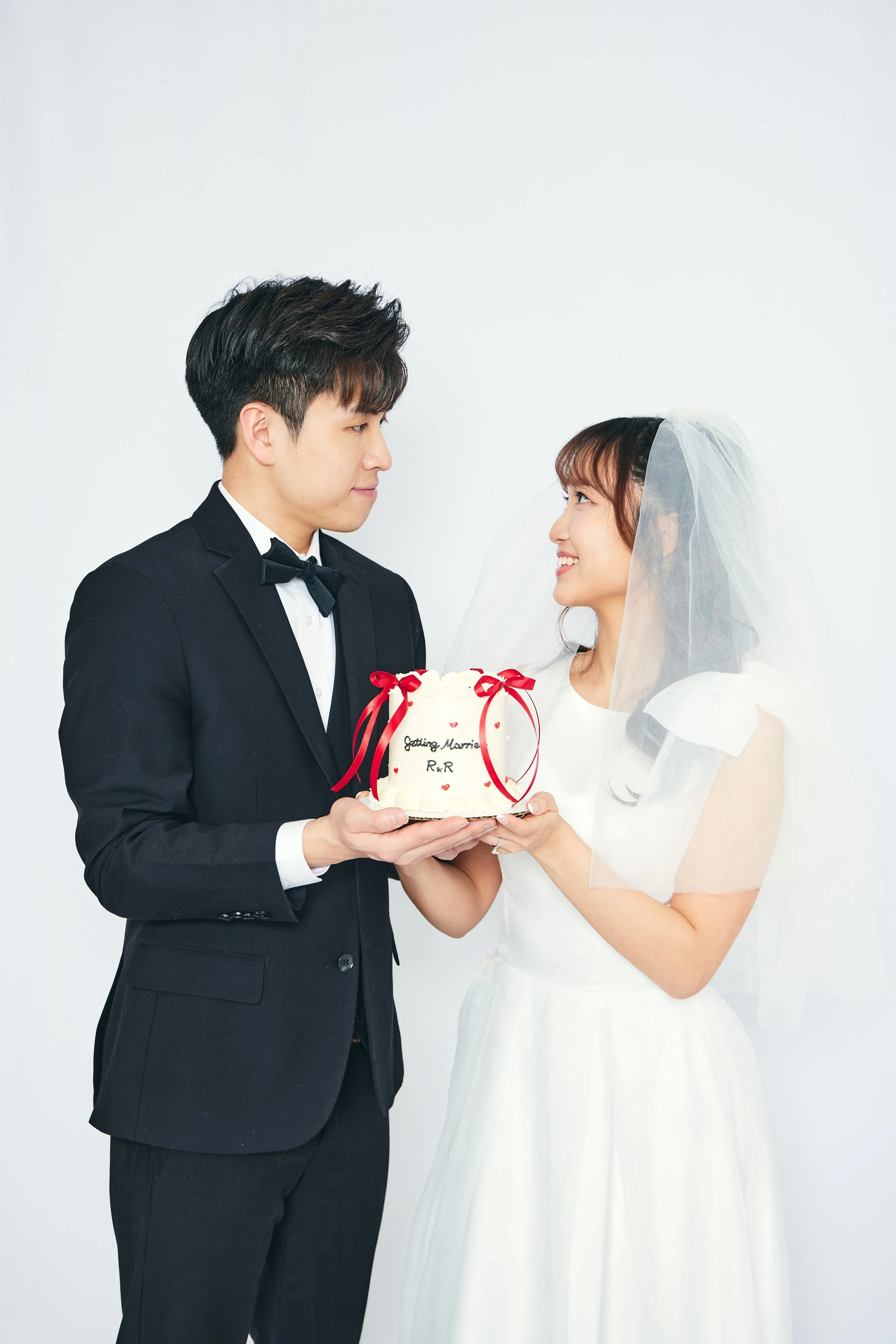 A bride and groom in wedding attire holding a white wedding cake with red ribbons and text that says, 'Getting Married R&R'.