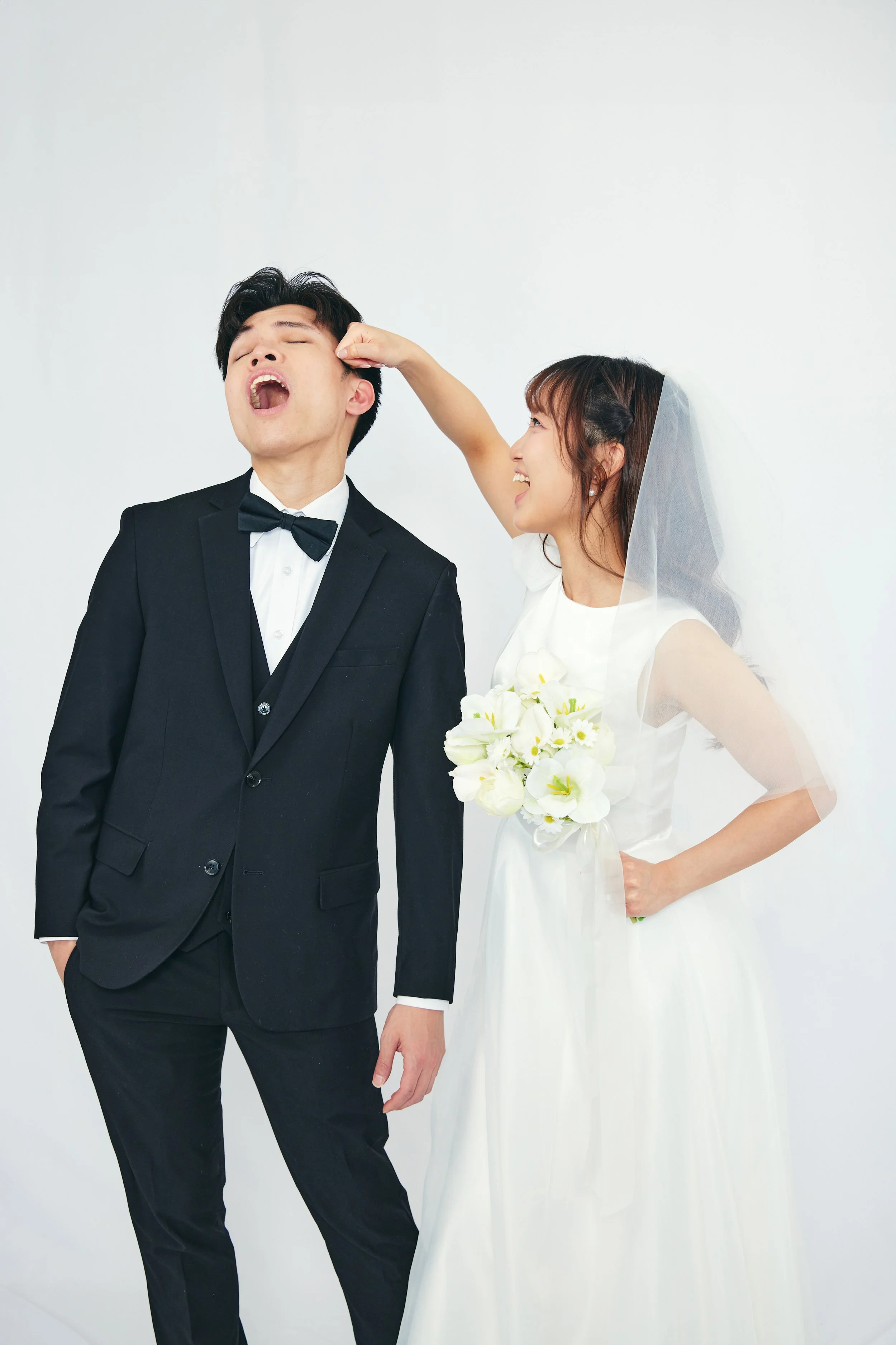 Bride playfully hitting groom's face with her fist during wedding photos as groom laughs with eyes closed, both in formal wedding attire, white background.