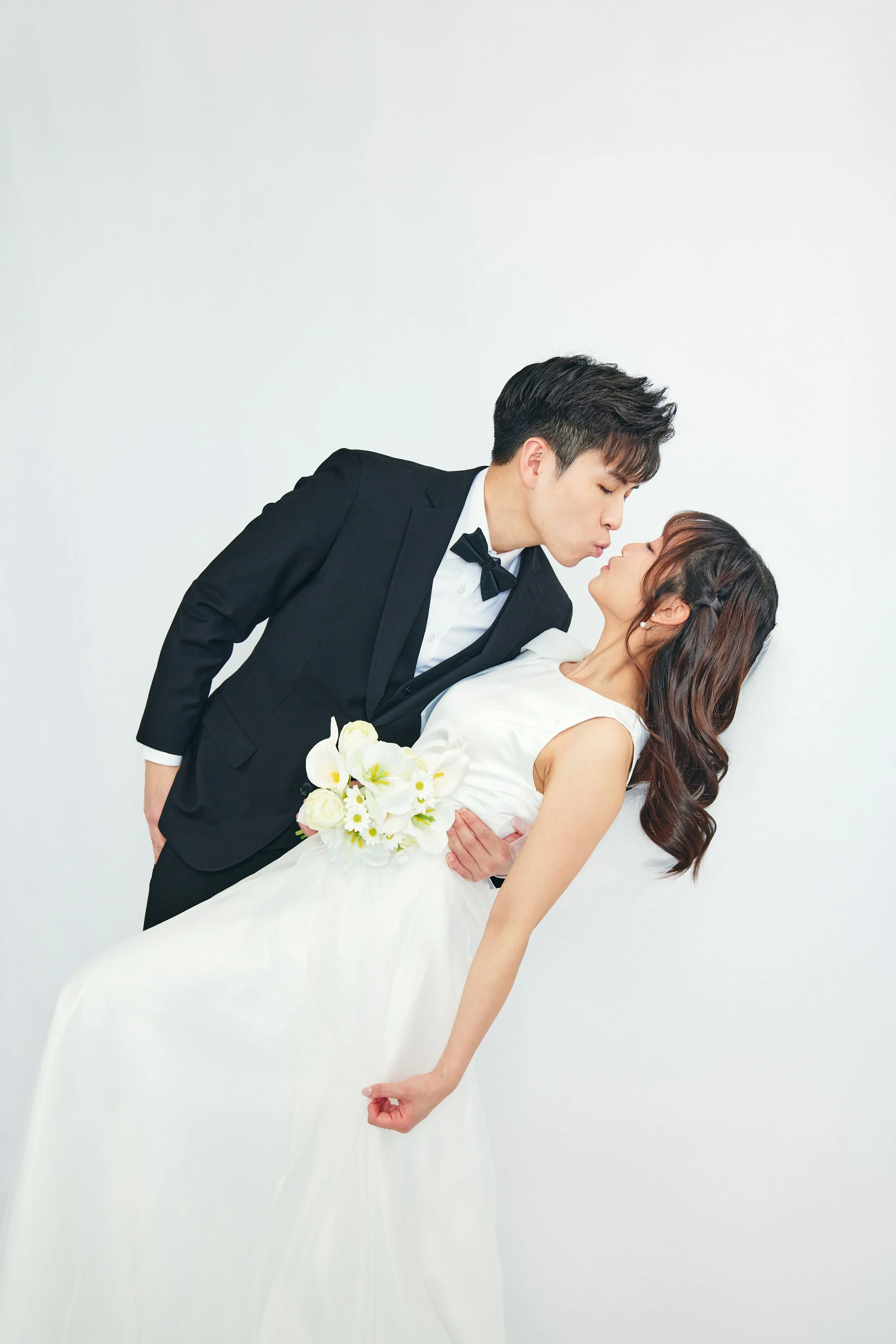 A groom in a black tuxedo leaning to kiss a bride in a white wedding gown, who is holding a bouquet of white flowers, against a plain white wall.