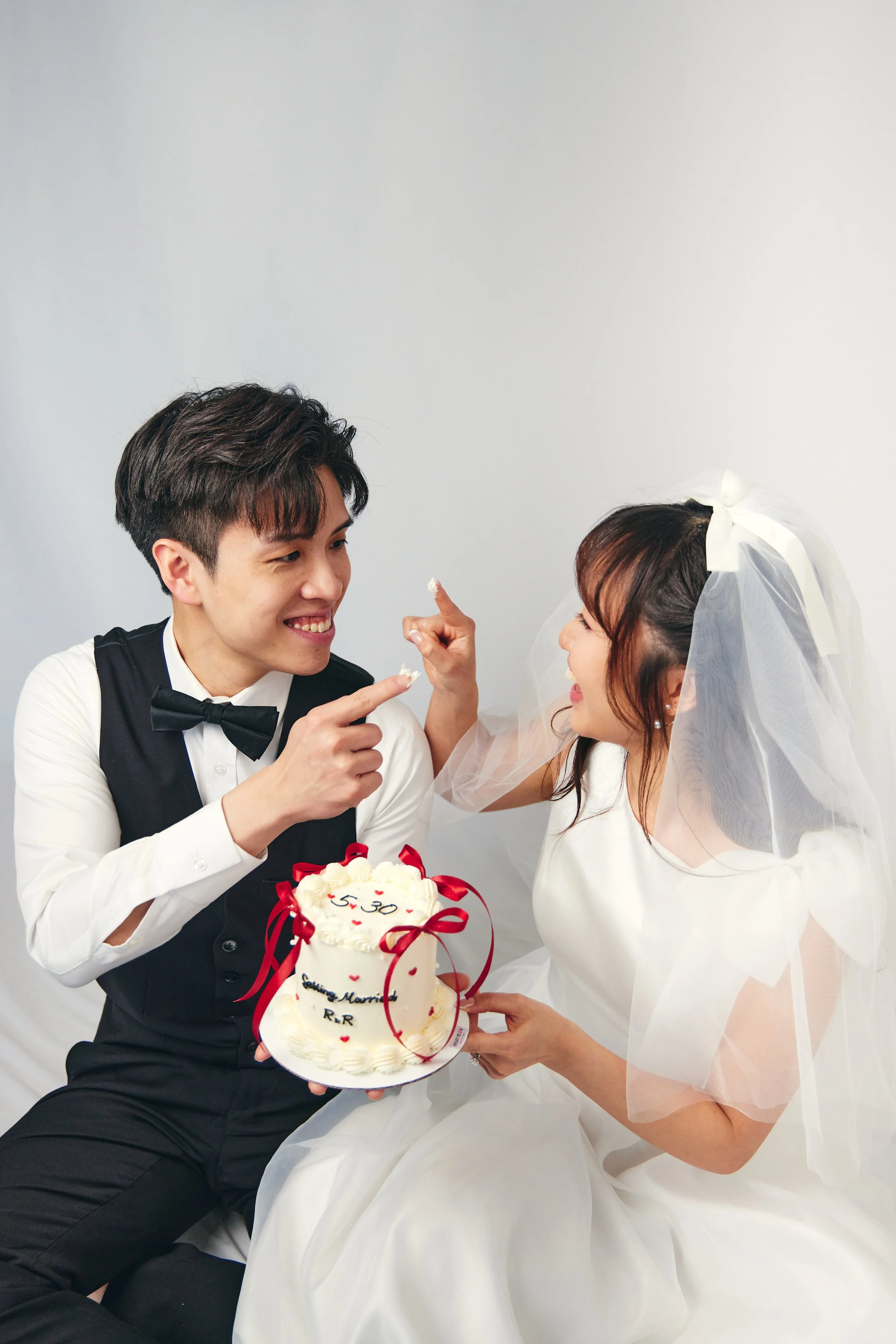 A young couple celebrating their wedding with a cake, the groom is smiling and pointing at the bride, who is wearing a wedding dress and veil, while holding the cake that says "5.30" and "happy married".