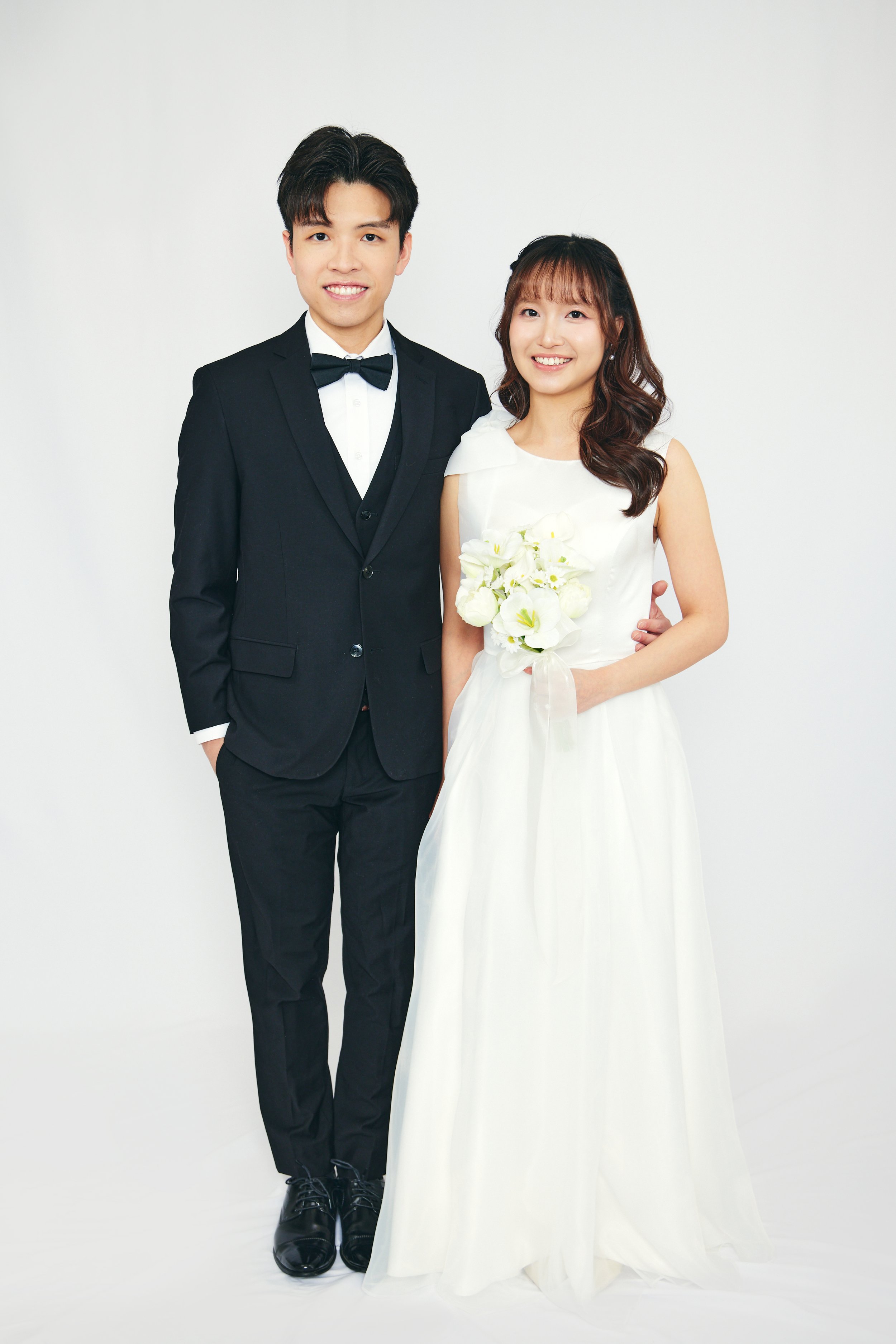 A newlywed couple dressed in wedding attire standing together against a plain white background, smiling for the camera. The groom is wearing a black tuxedo with a bow tie and the bride is wearing a white wedding dress holding a bouquet of white flowe