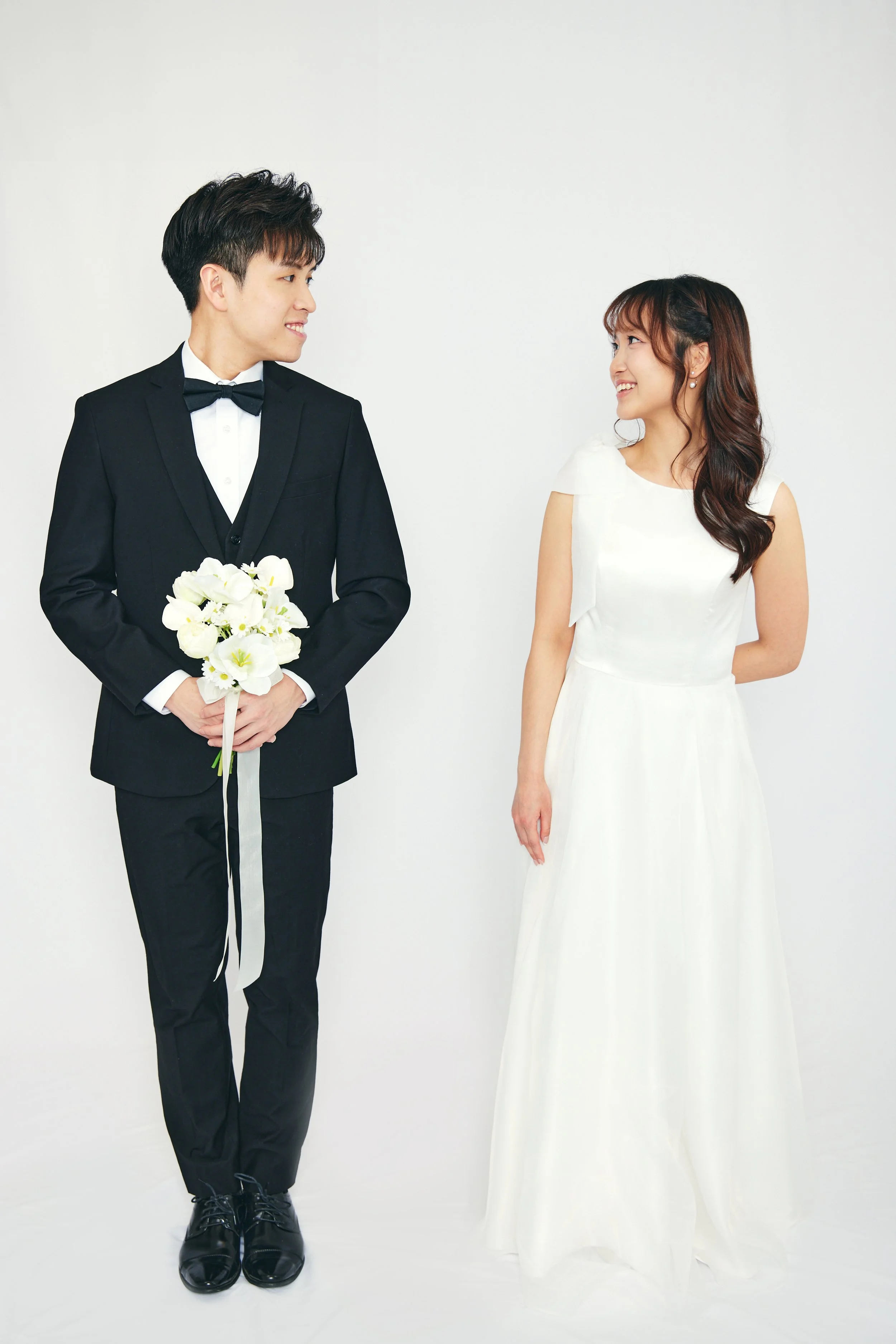 A couple dressed in wedding attire, the groom in a black tuxedo holding a white bouquet, and the bride in a white gown, smiling at each other against a plain white background.