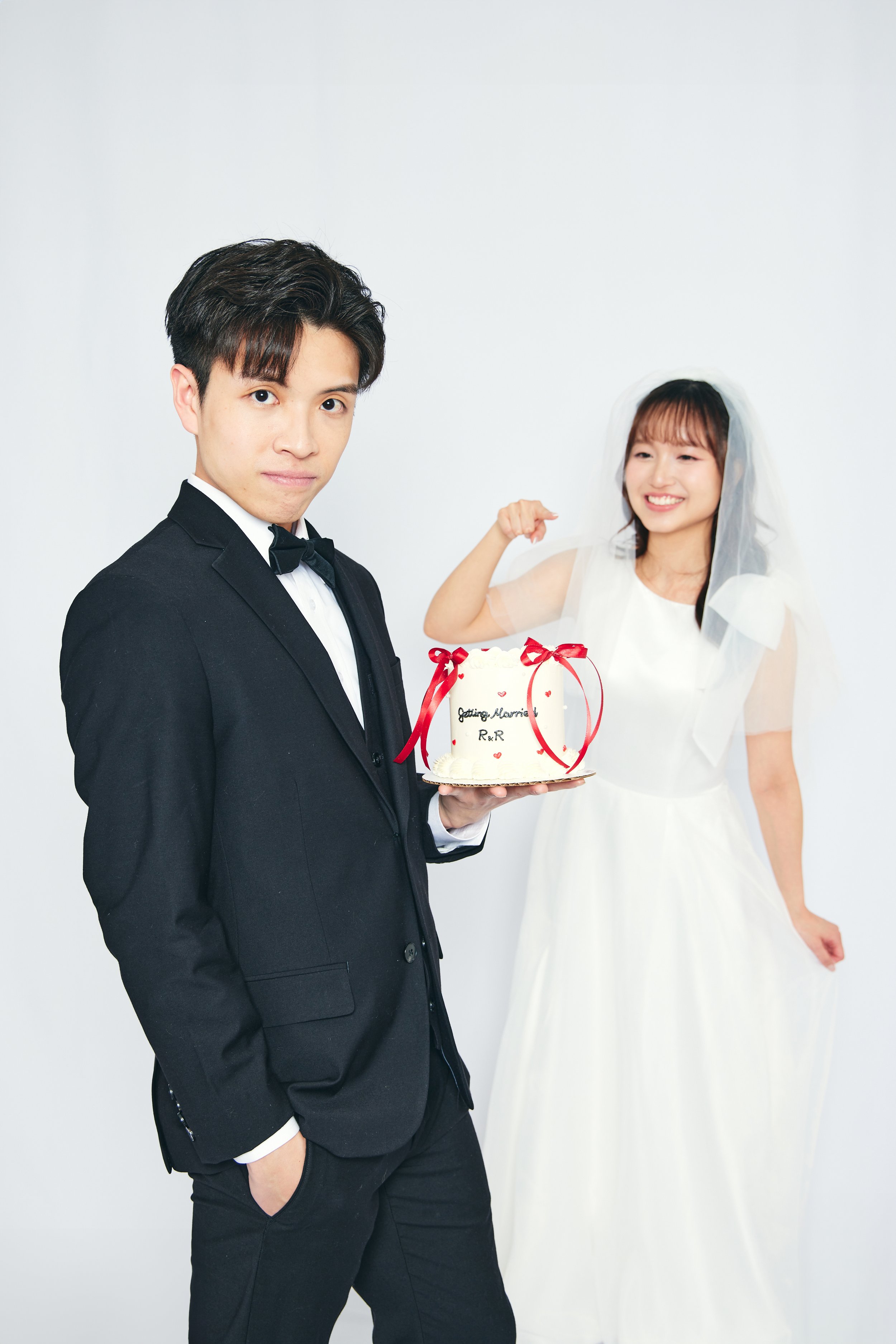 A bride and groom at their wedding, with the groom holding a wedding cake decorated with red ribbons and small hearts, and the bride smiling in the background.