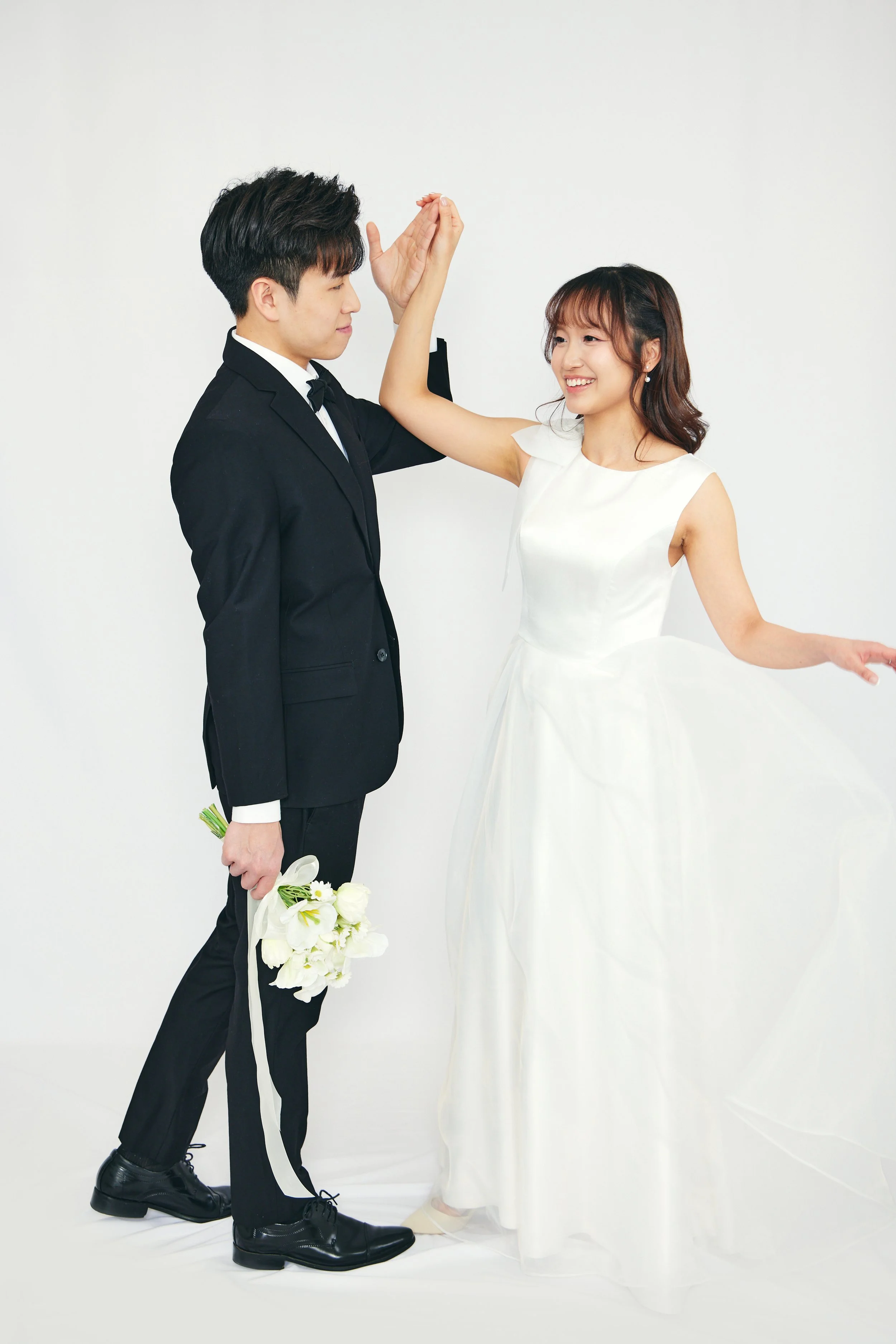 A bride and groom dancing together, with the groom holding a bouquet of flowers, in a plain white background.