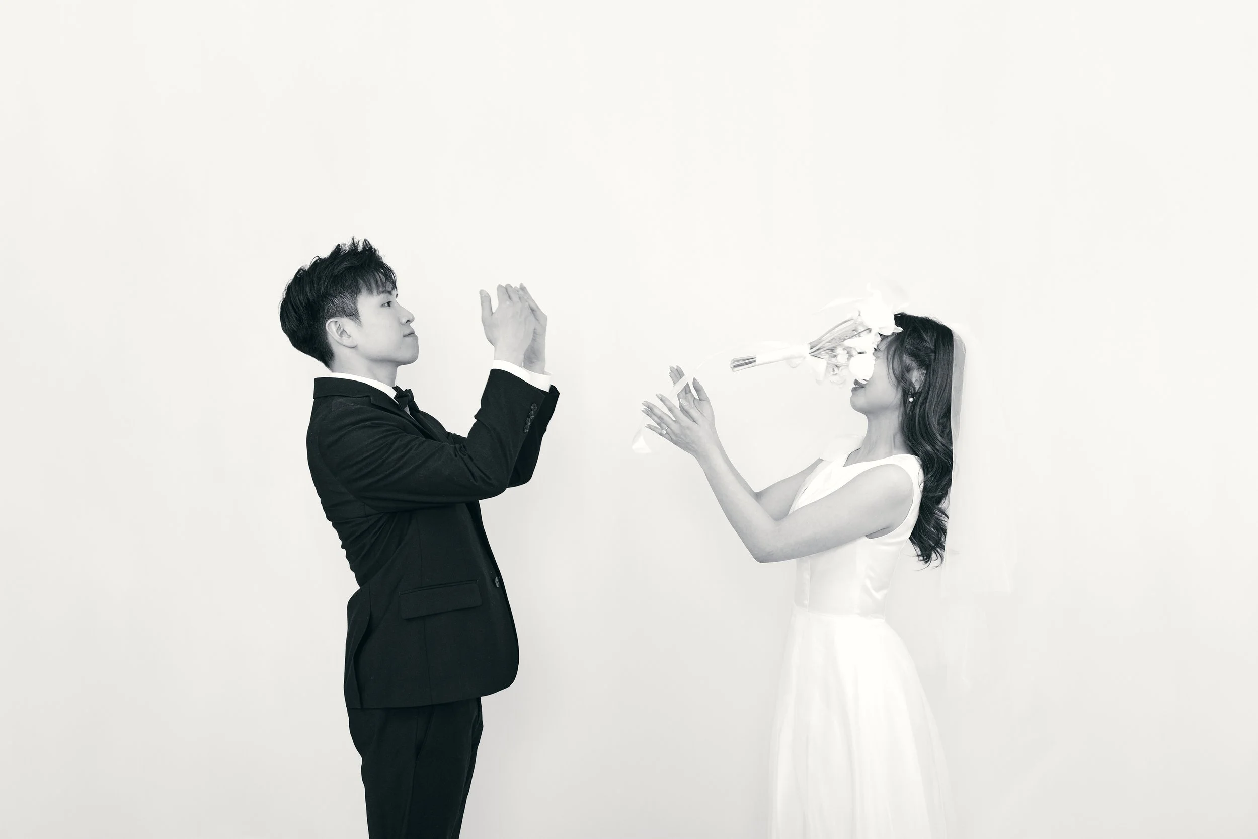 A black-and-white photo of a wedding scene with a man in a suit and a woman in a wedding dress. The woman is blowing confetti out of a party blower, and the man is clapping.