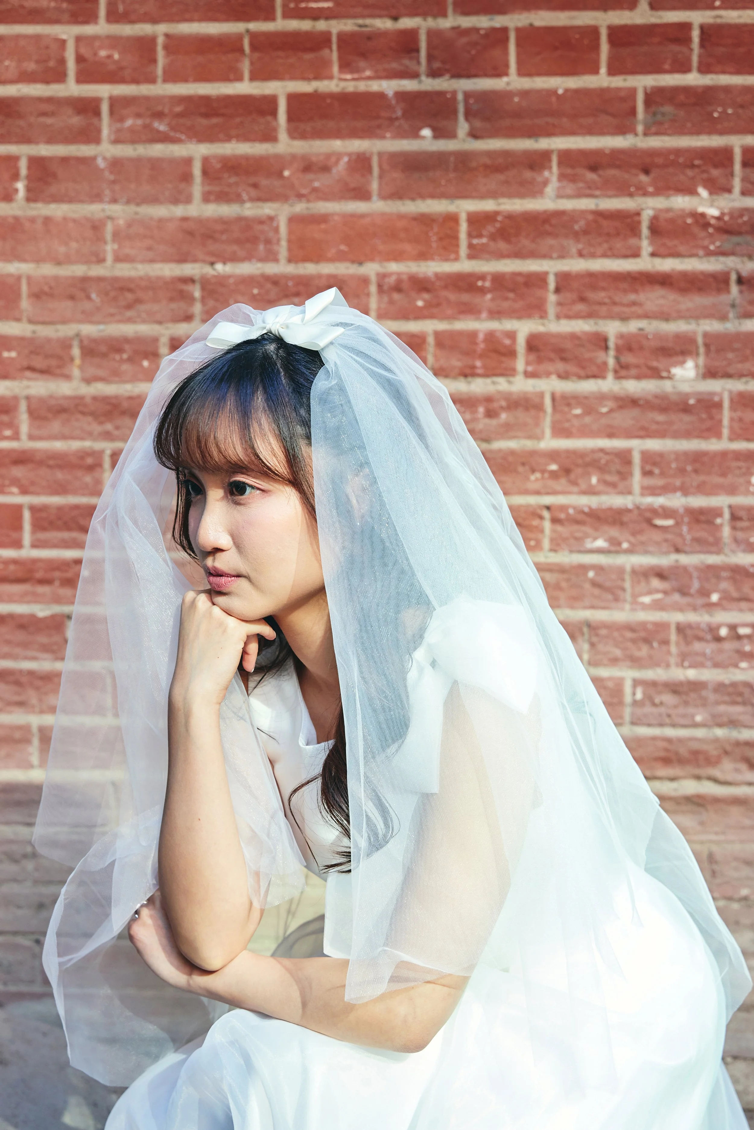 A woman in a white dress with a veil and bow, sitting with her chin resting on her hand against a brick wall background.