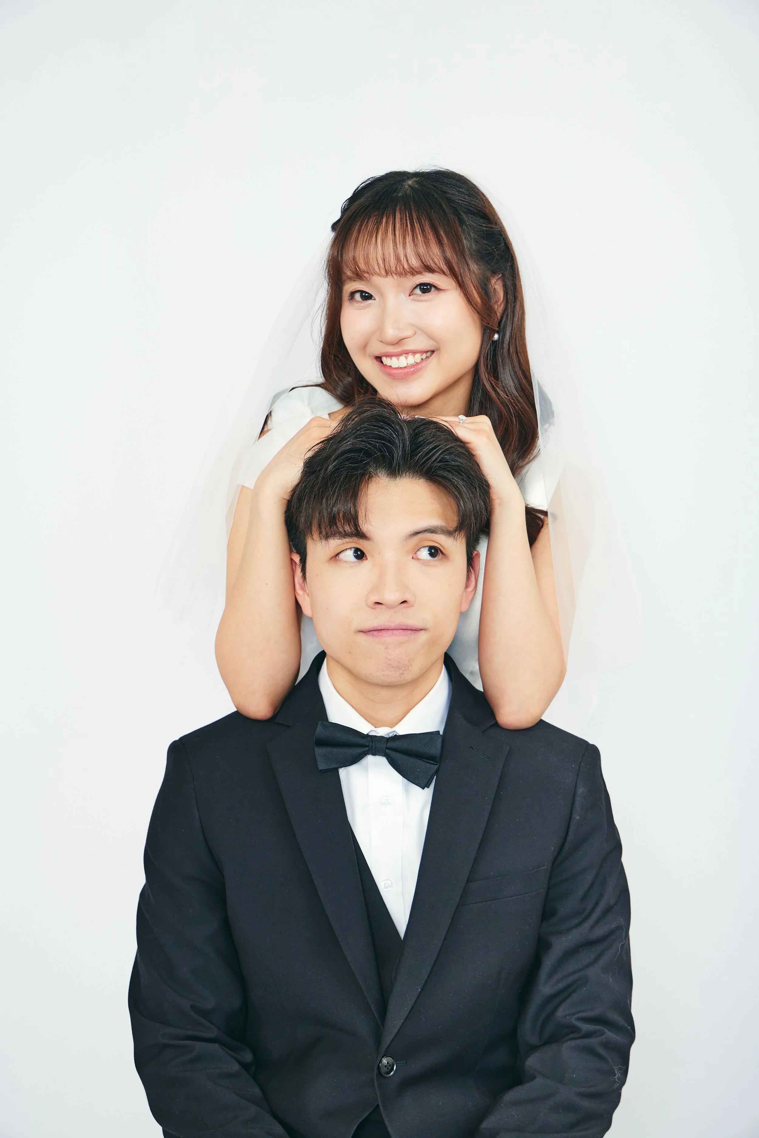 A happy young woman in a wedding dress and veil making a playful pose with her head resting on the young man's head, who is dressed in a black tuxedo with a bowtie, against a plain white background.