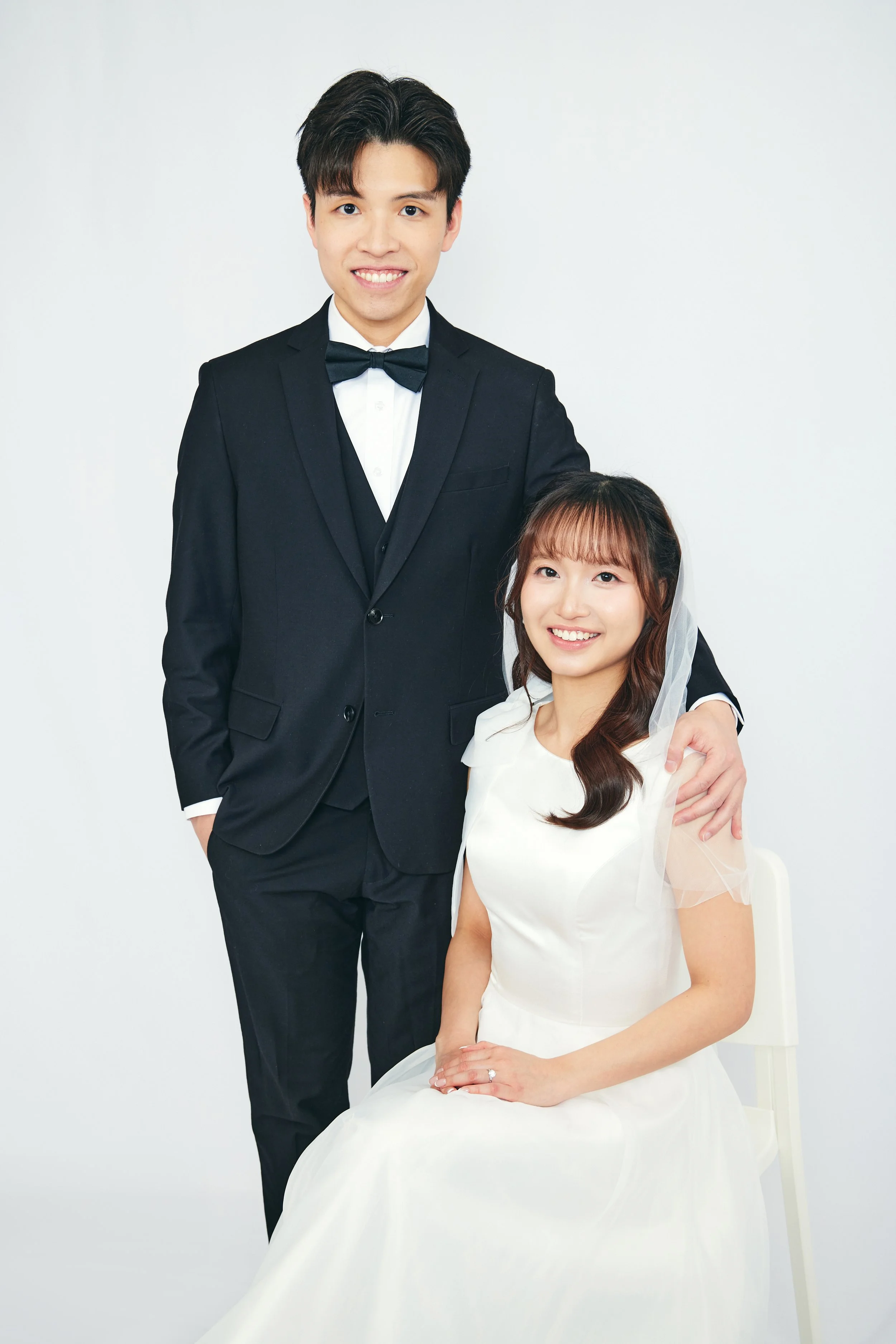 A man in a black tuxedo and a woman in a white dress, sitting and standing together, smiling at the camera against a plain white background.