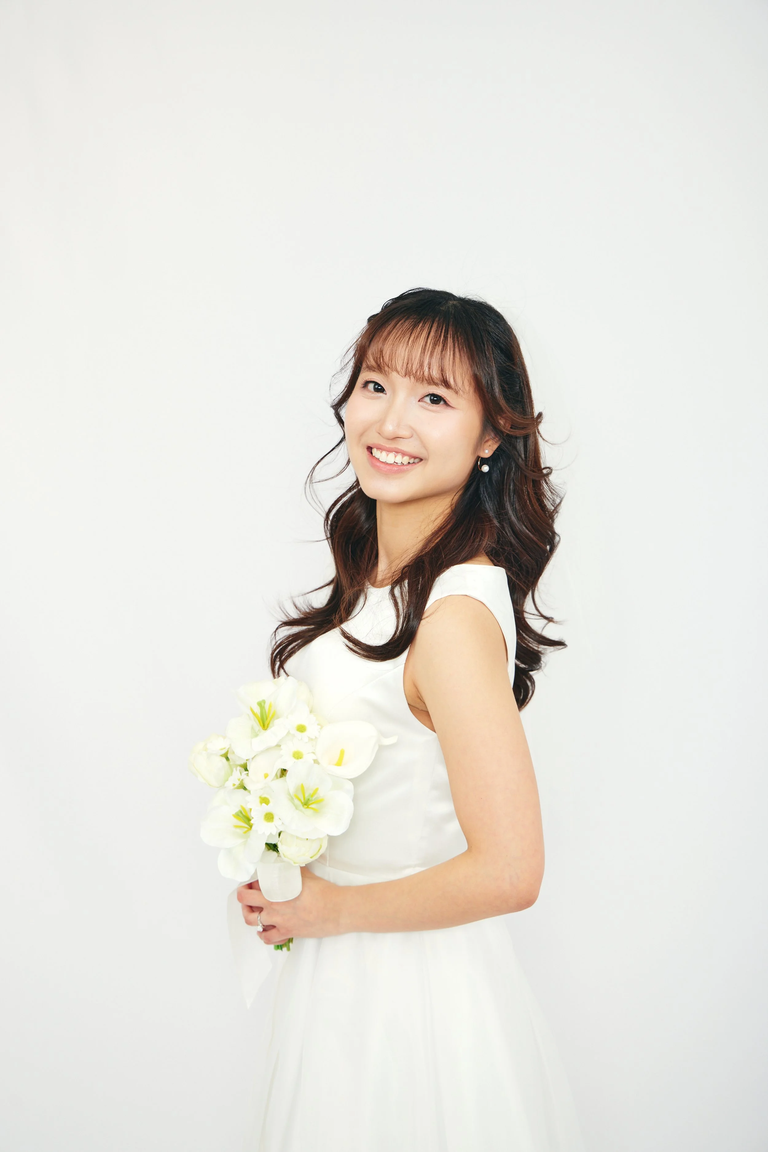Smiling young woman in a white dress holding a bouquet of white flowers, standing against a plain white background.