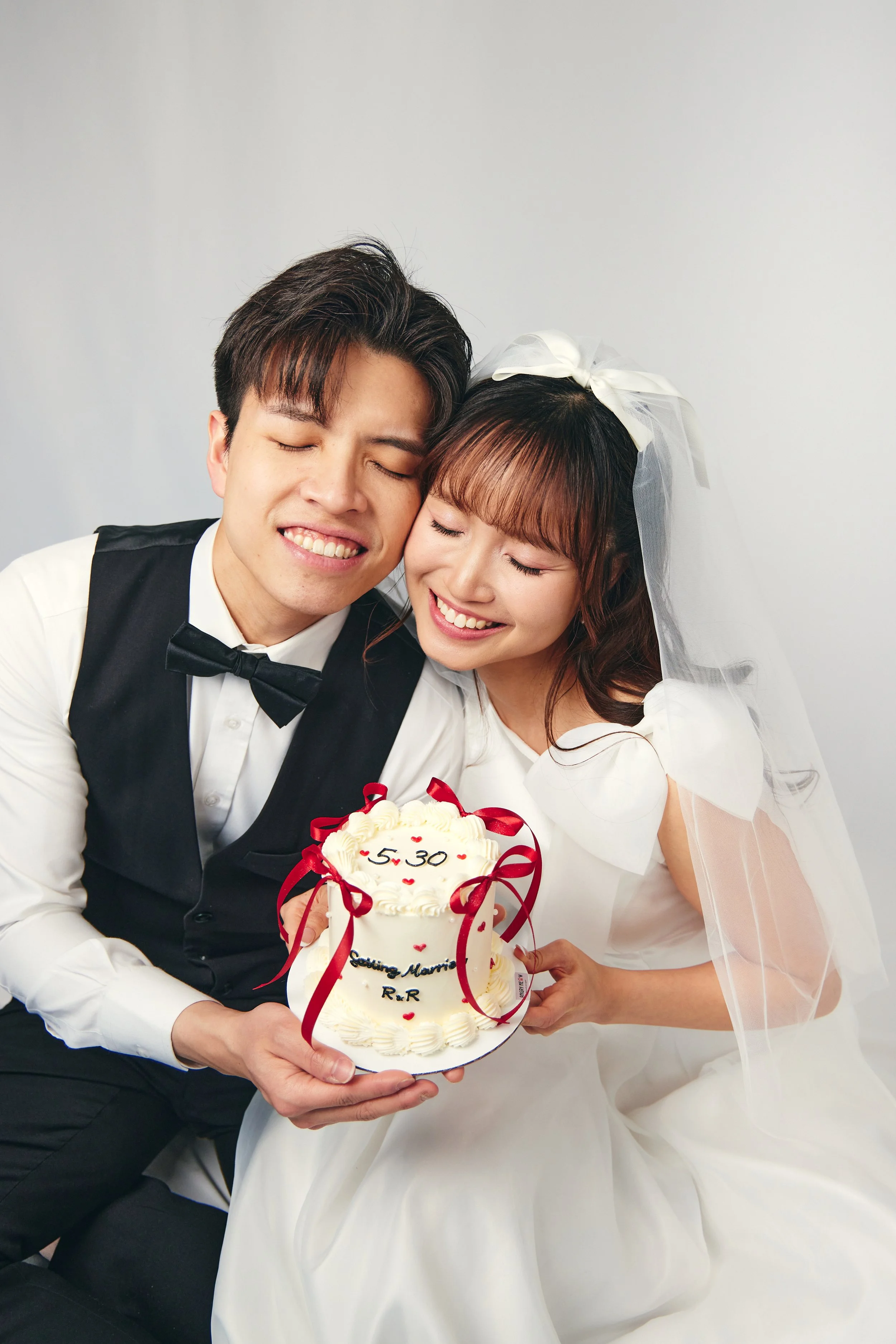 A happy bride and groom in wedding attire holding a wedding cake with the date 5-30 decorated on it, smiling and pressing their heads together.