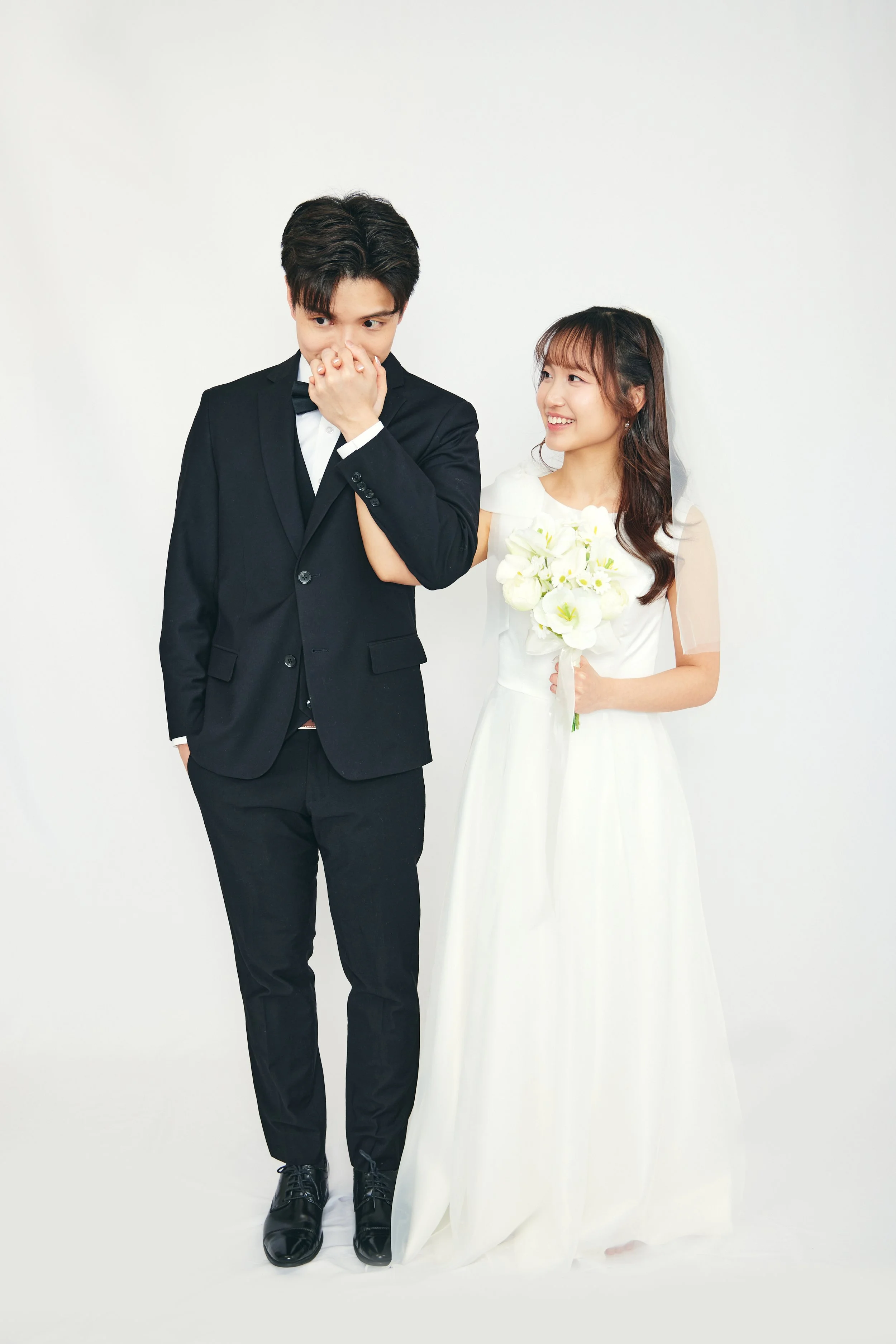 A bride in a white wedding dress holding a bouquet, smiling at a groom in a black suit who cover his mouth with his hand, both standing against a white background.