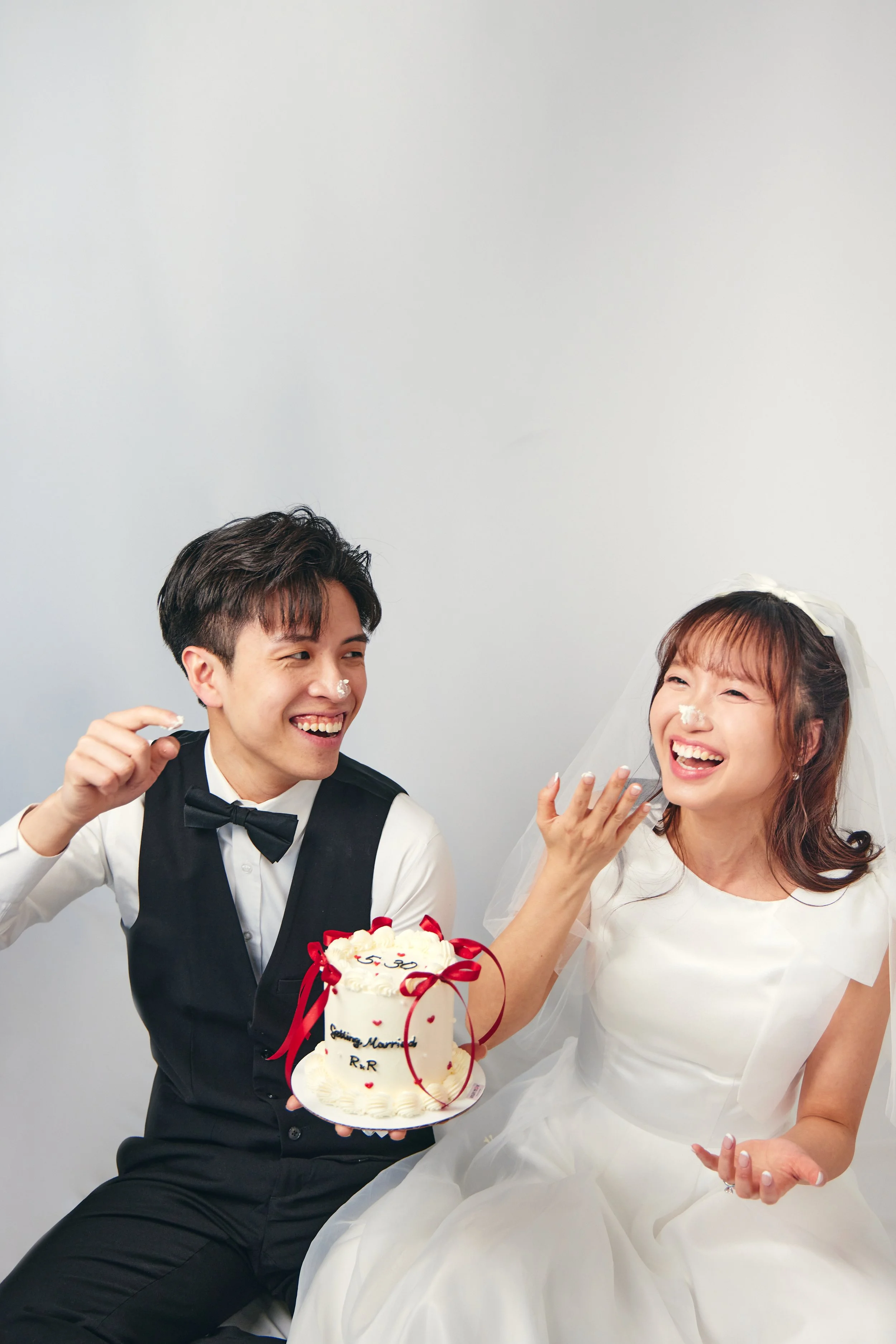 A happy couple at their wedding, sharing a cake and smiling, with white frosting and red ribbons on the cake.