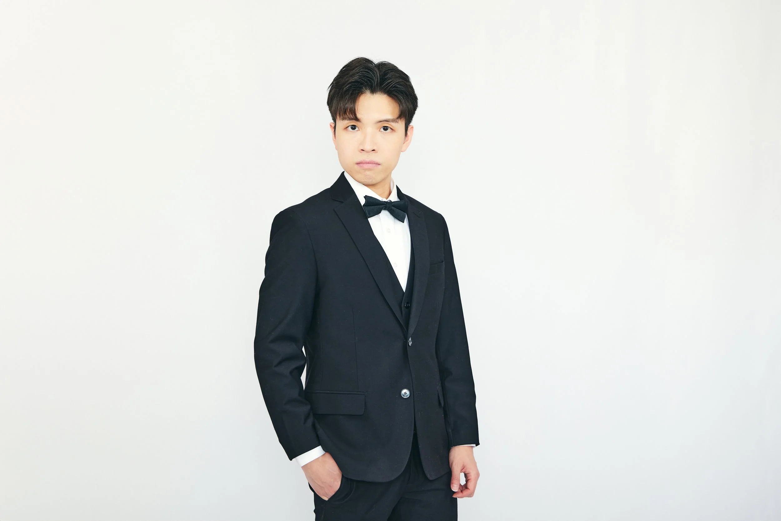 Young man in a black tuxedo and bow tie standing against a plain white background.