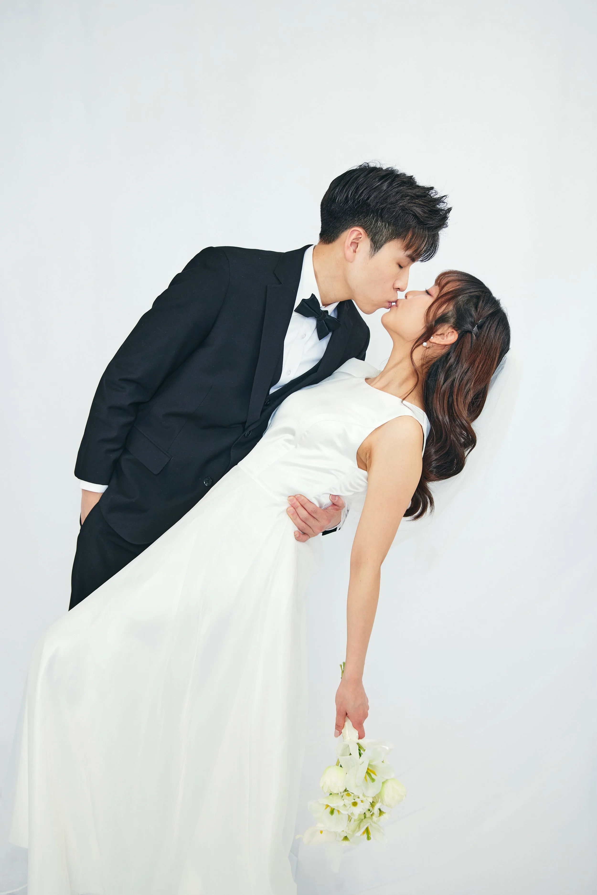 A couple dressed in wedding attire sharing a kiss, with the groom in a black tuxedo and the bride in a white wedding dress holding a bouquet of white flowers.