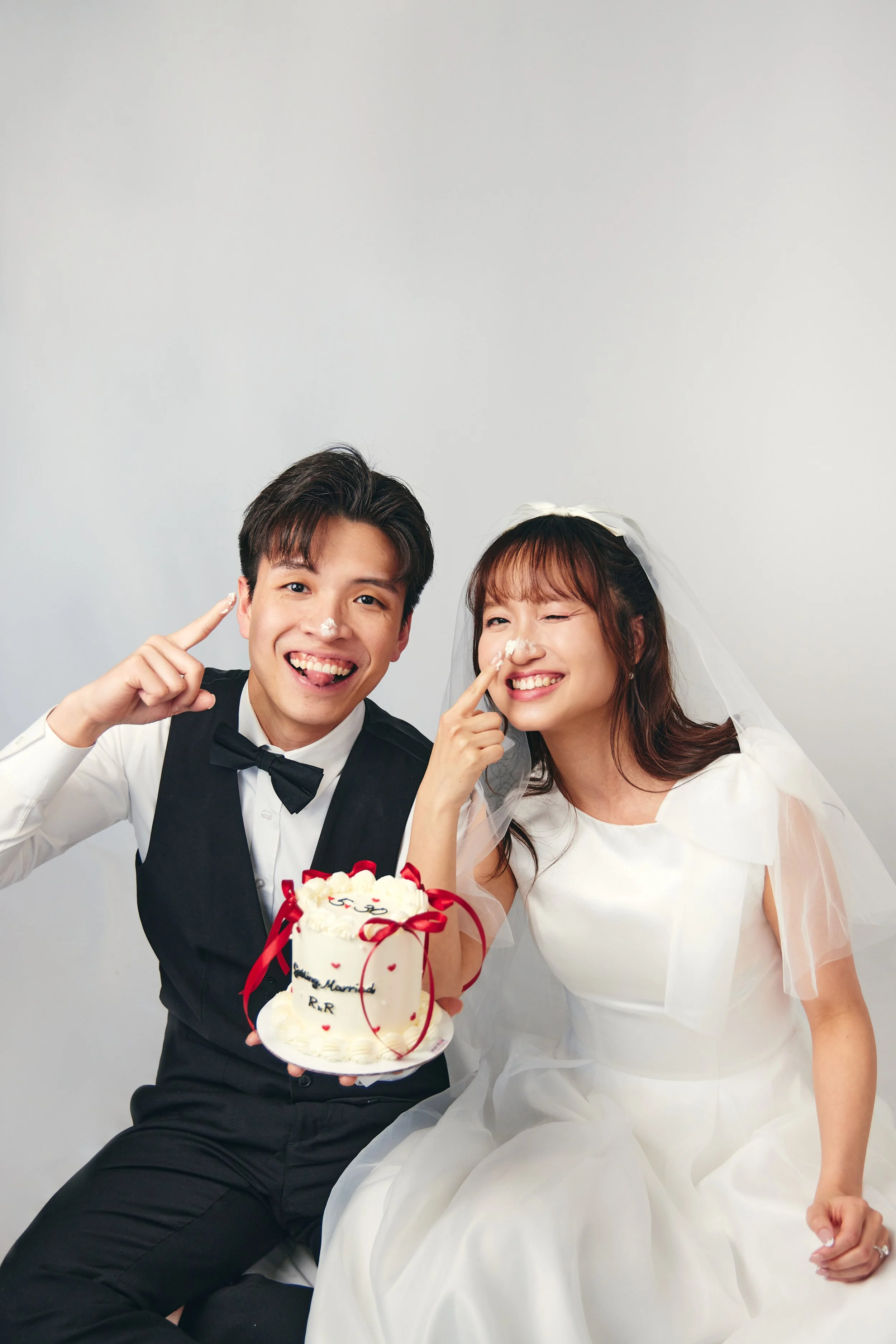Happy newlywed couple in wedding attire celebrate with cake, smiling and pointing at their noses, against a plain white background.