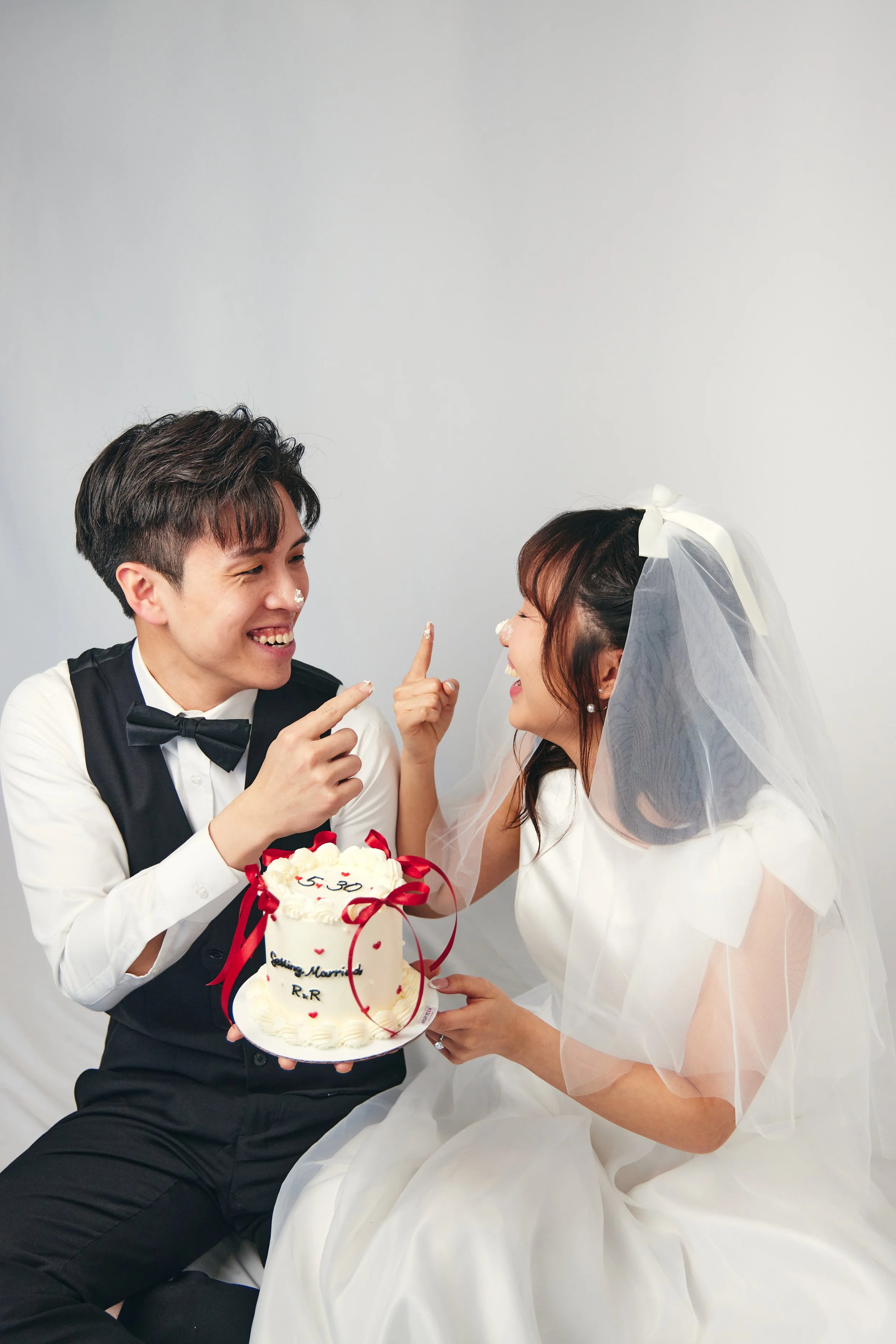 A bride and groom laughing while holding a wedding cake, with the groom pointing at the bride's nose which has cake on it.