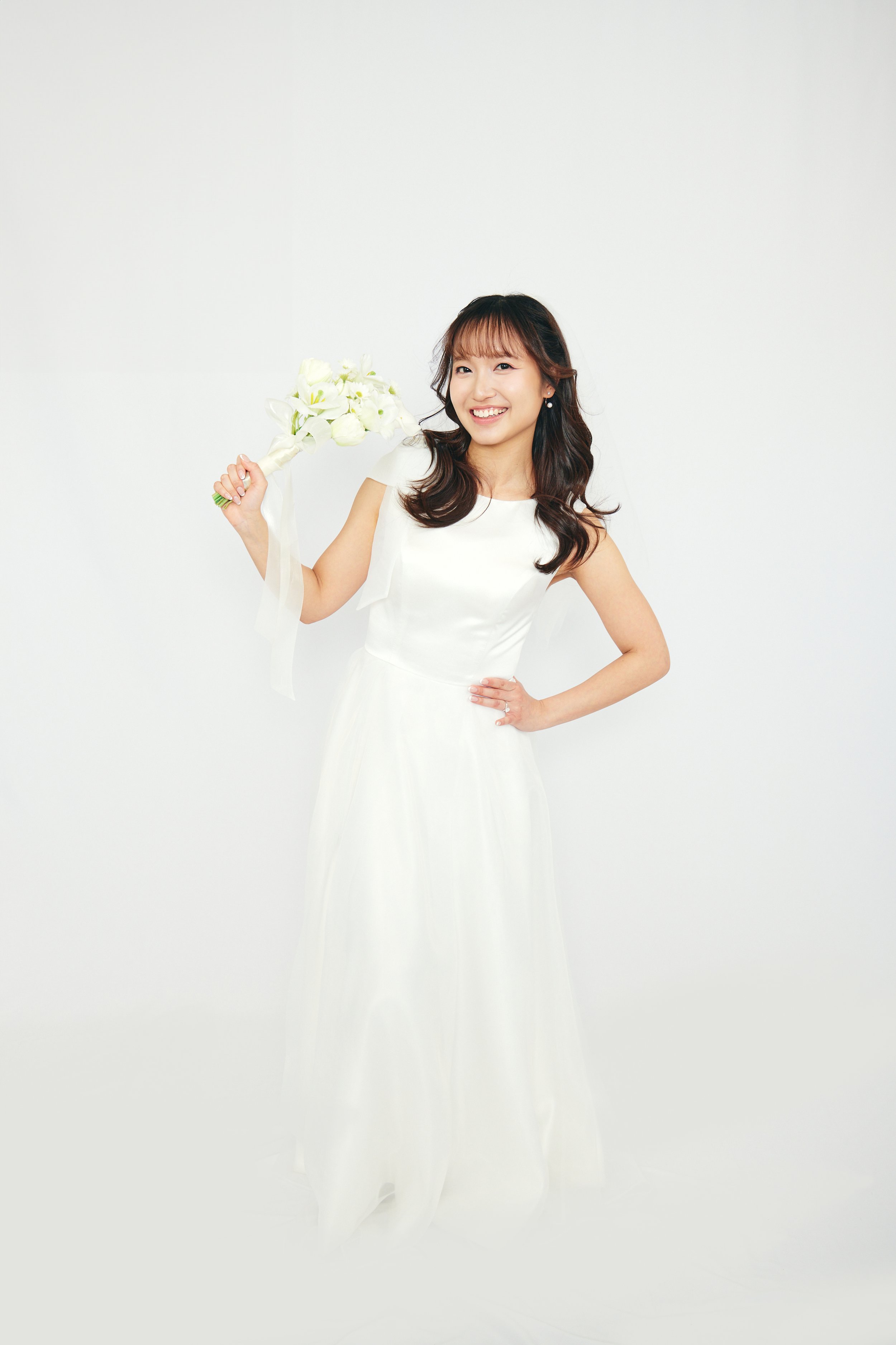 A woman in a white dress holding a bouquet of white flowers, smiling against a plain white background.