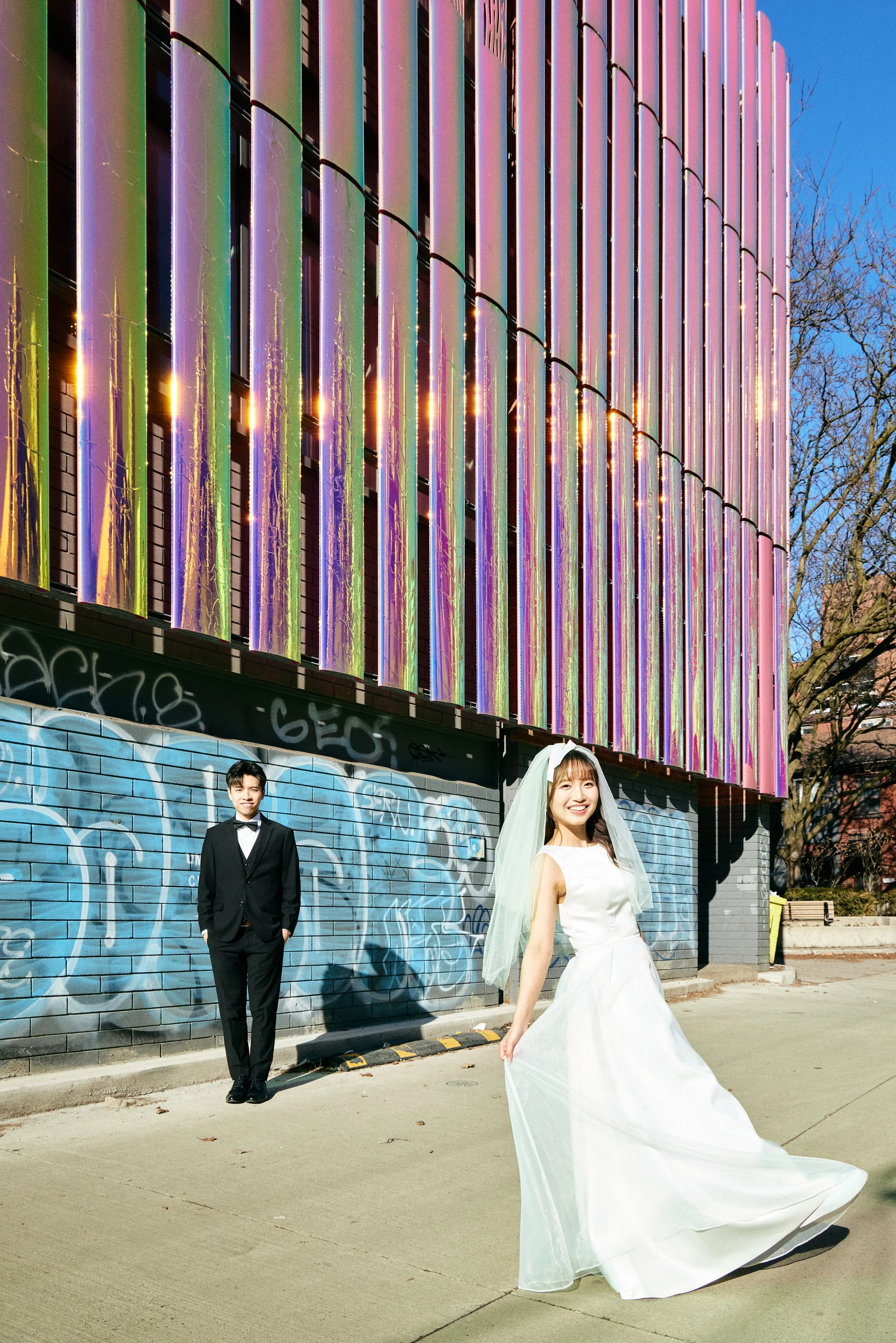 A bride in a white wedding dress and veil smiling, holding her dress, standing on a sidewalk. A groom in a black tuxedo and bow tie stands nearby. Behind them is a graffiti-covered wall and a building with colorful vertical neon lights or artwork on 