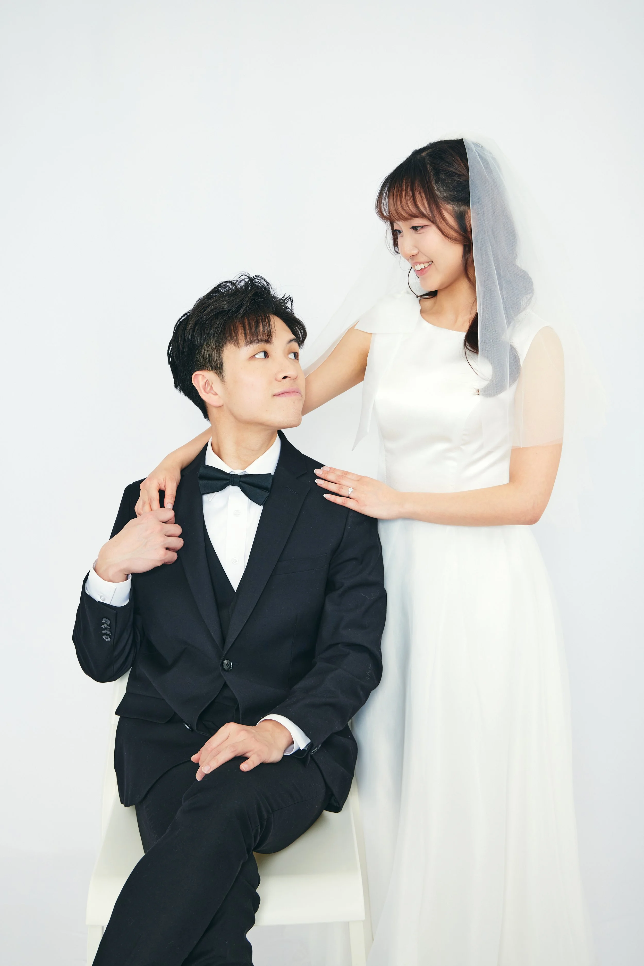 A groom in a black tuxedo sitting and looking up at a smiling bride in a white wedding dress and veil who is standing beside him with her hand on his shoulder.