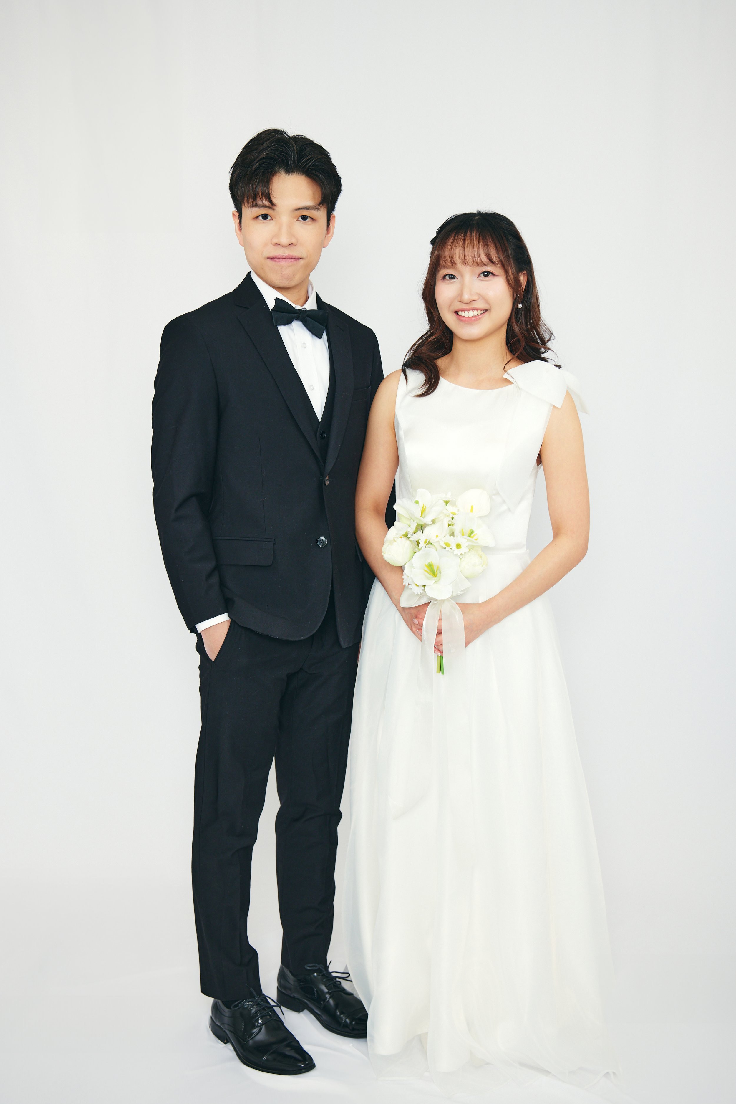 A bride and groom standing together in wedding attire against a plain white background. The groom is wearing a black tuxedo with a bow tie, and the bride is wearing a white wedding dress holding a bouquet of white flowers.