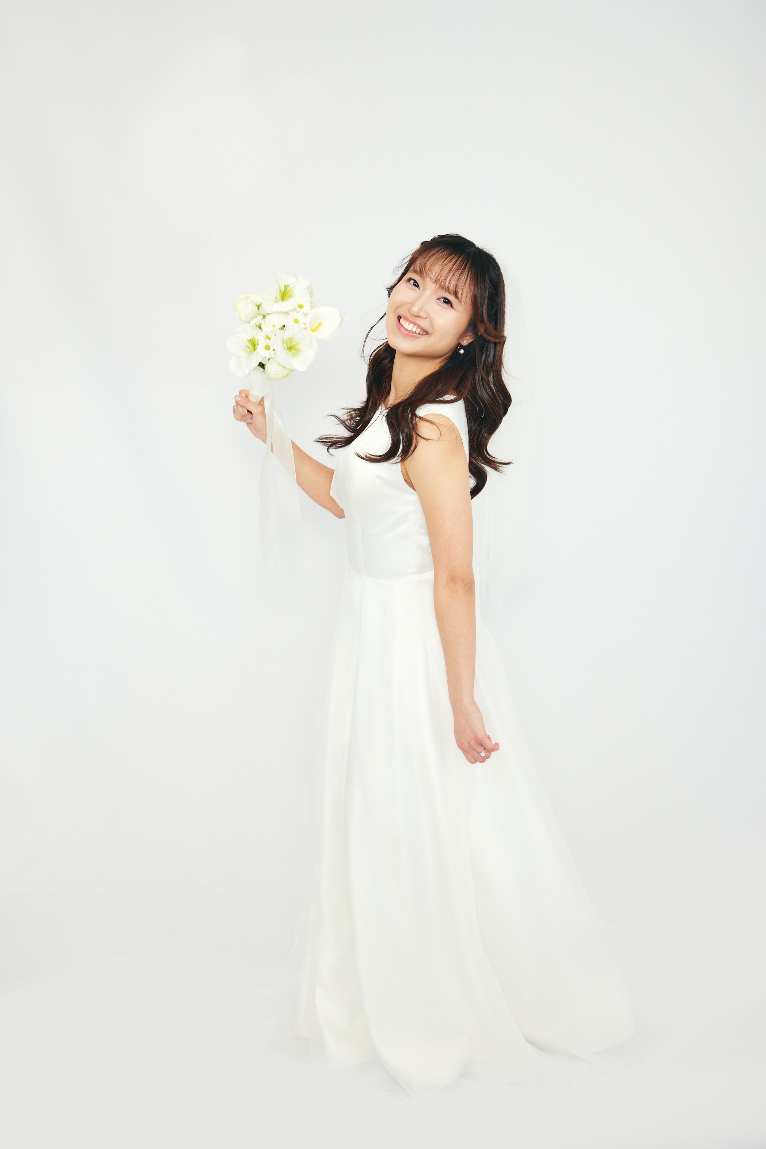 A woman in a white dress holding a bouquet of white flowers, smiling and standing against a plain white background.
