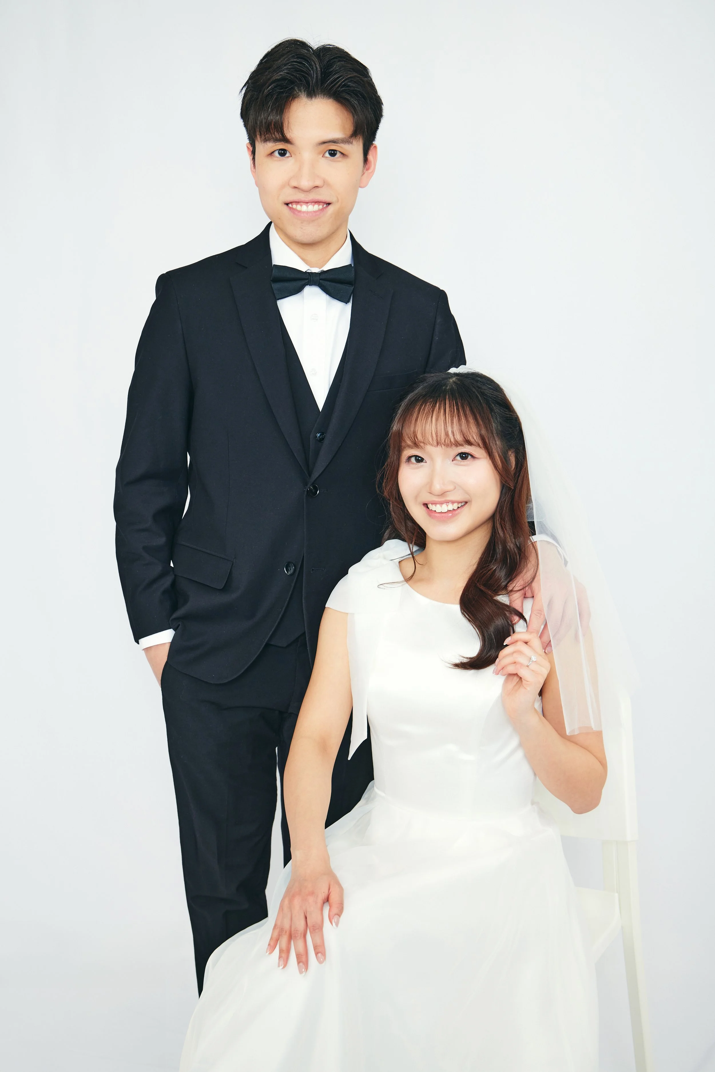 A happy couple dressed in wedding attire, the man in a black tuxedo and the woman in a white wedding dress, in a studio with a white background.