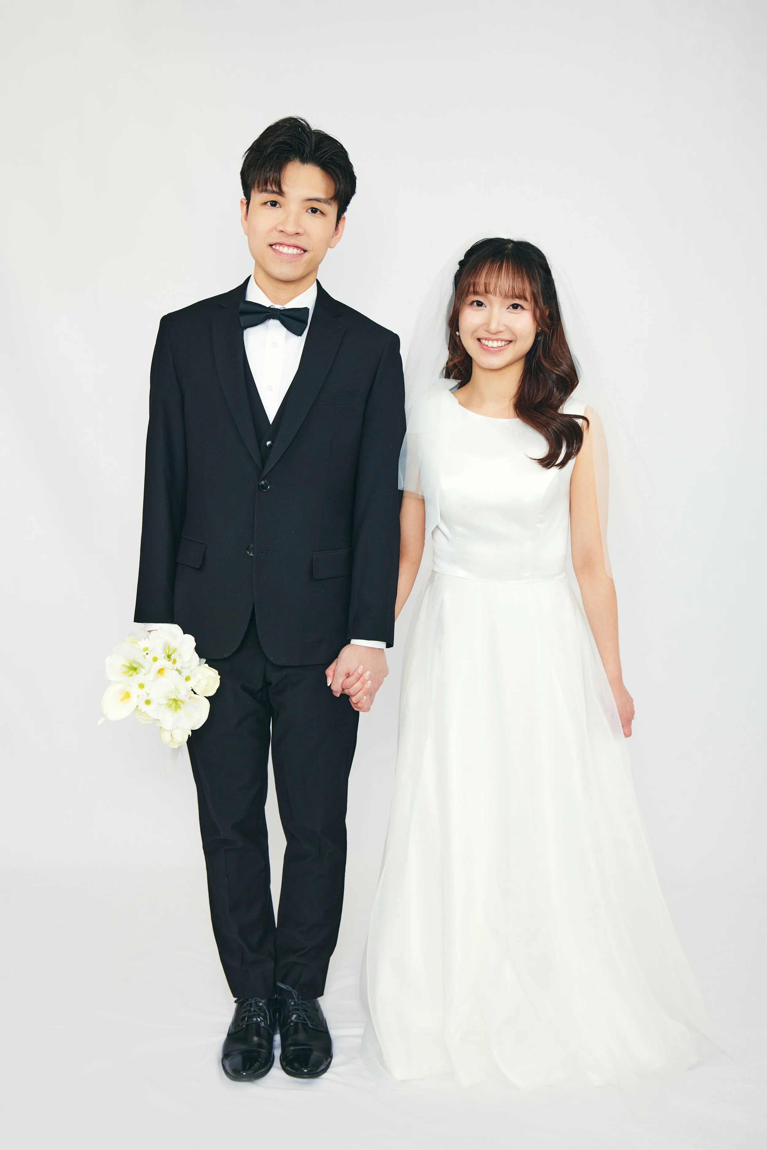 A smiling bride and groom holding hands, dressed in wedding attire, in front of a plain white background.