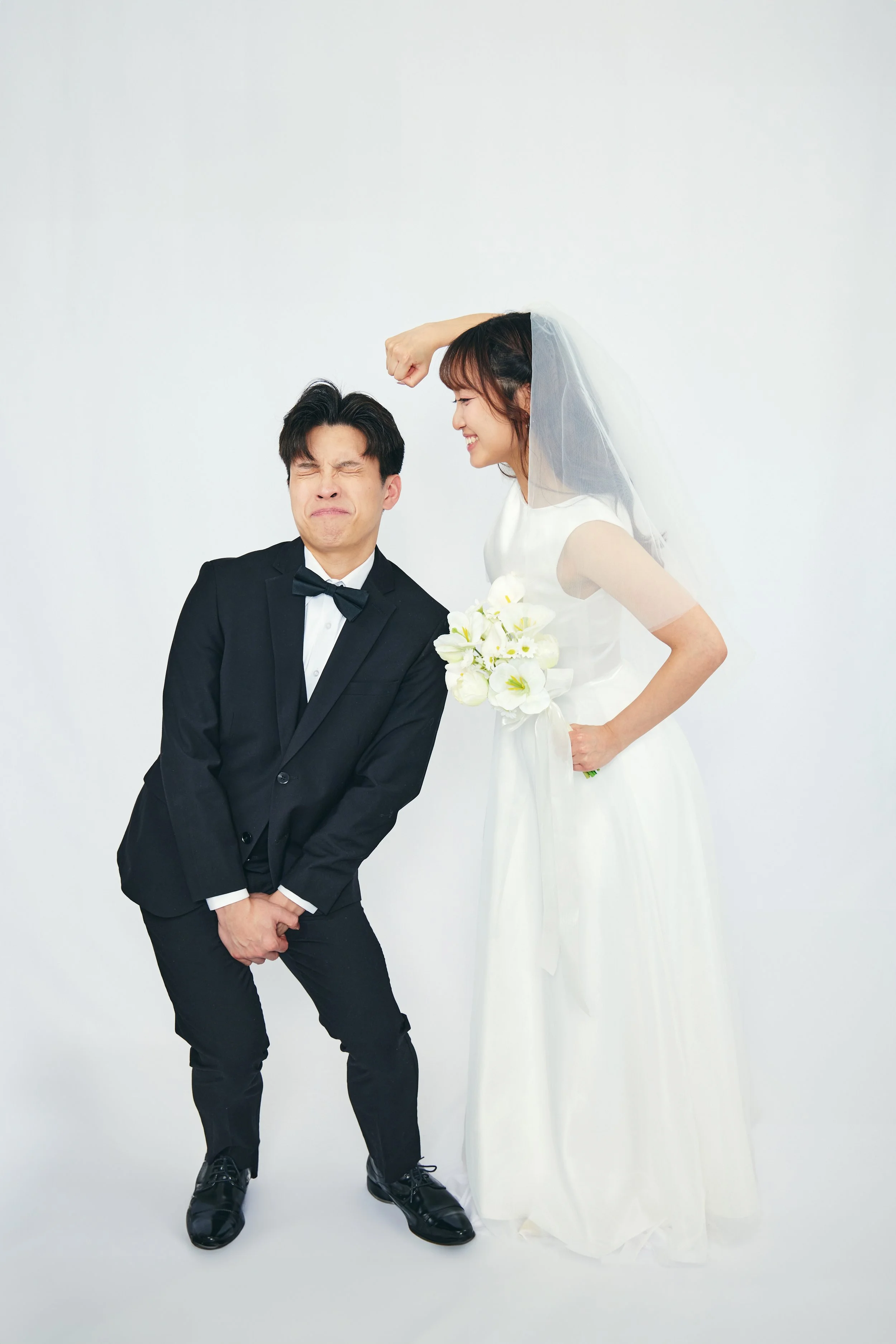 A bride in a white wedding dress holding a bouquet of flowers smiling and playfully flexing her arm towards a groom in a black tuxedo making a funny face.