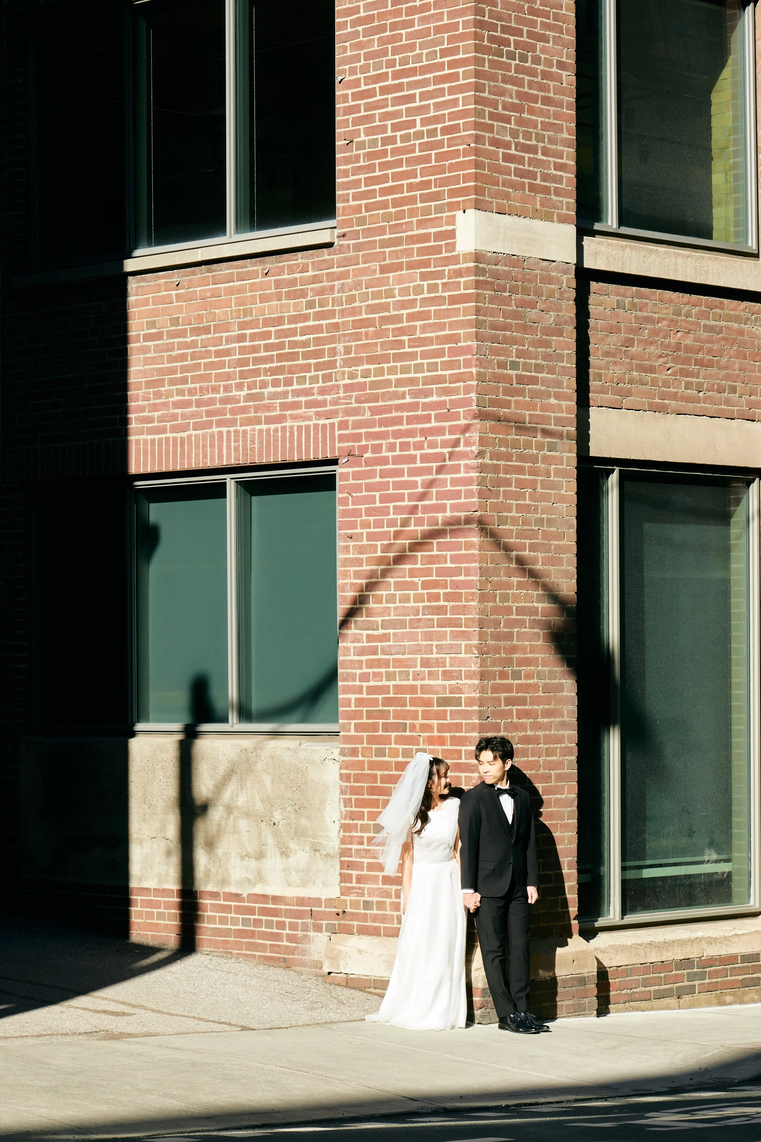A bride and groom in wedding attire standing outside against a brick building wall, holding hands and looking at each other.