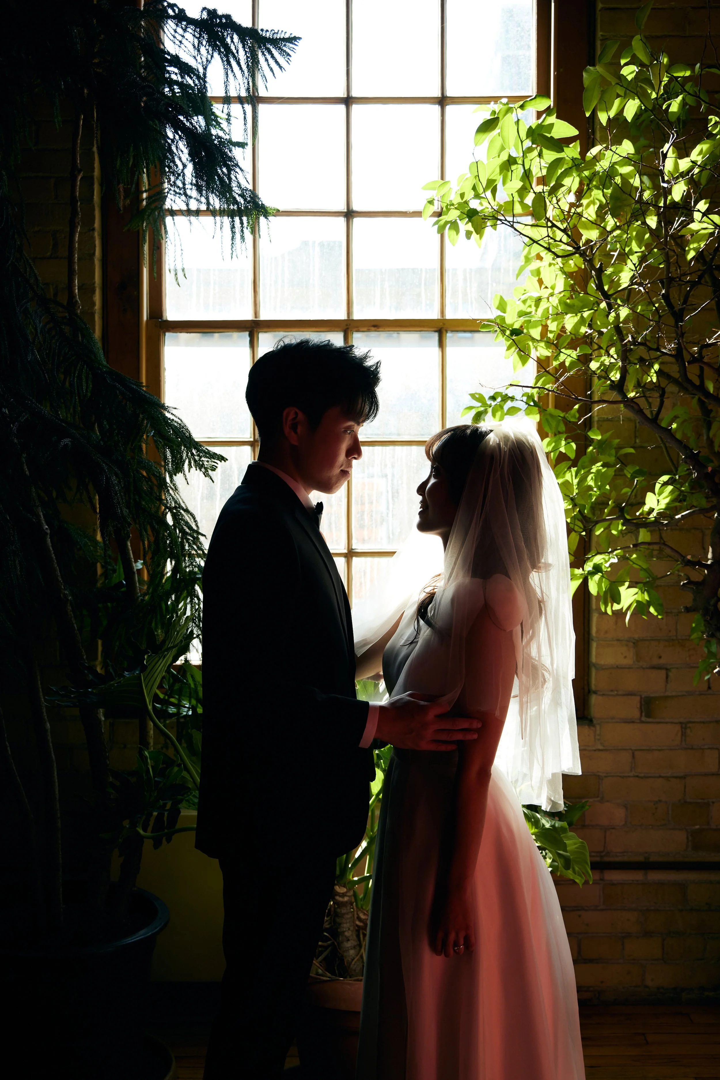 A bride and groom standing close to each other indoors near a large window with natural light, surrounded by green plants, with the groom holding the bride's waist.
