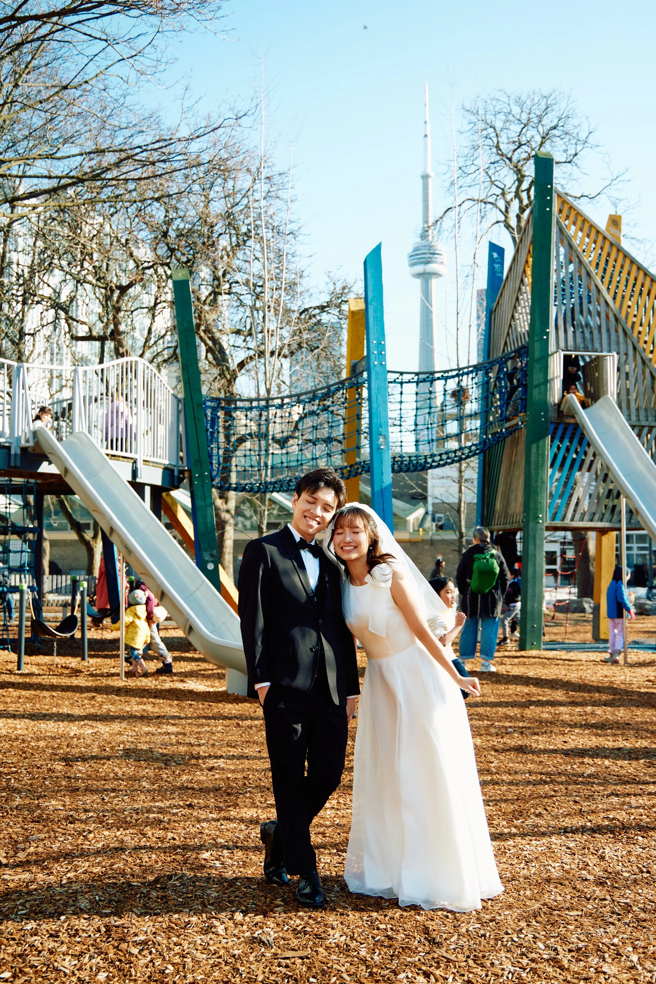 A newlywed couple in wedding attire smiling and leaning towards each other in a playground with slides and climbing structures, with the CN Tower visible in the background on a clear day.