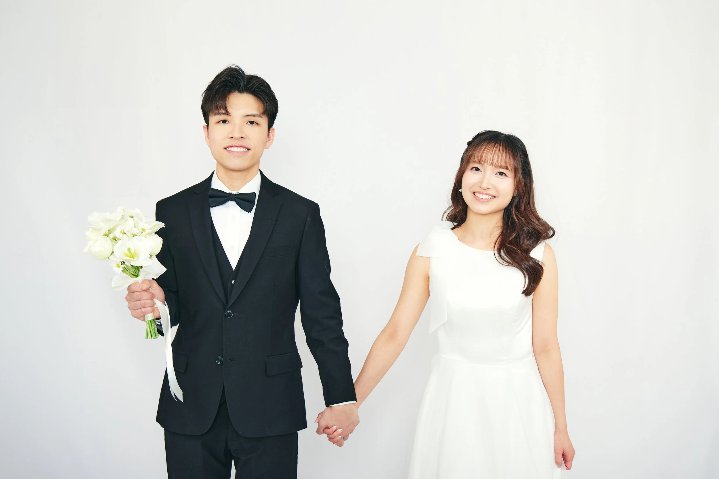A happy couple dressed in wedding attire holding hands against a plain white background.